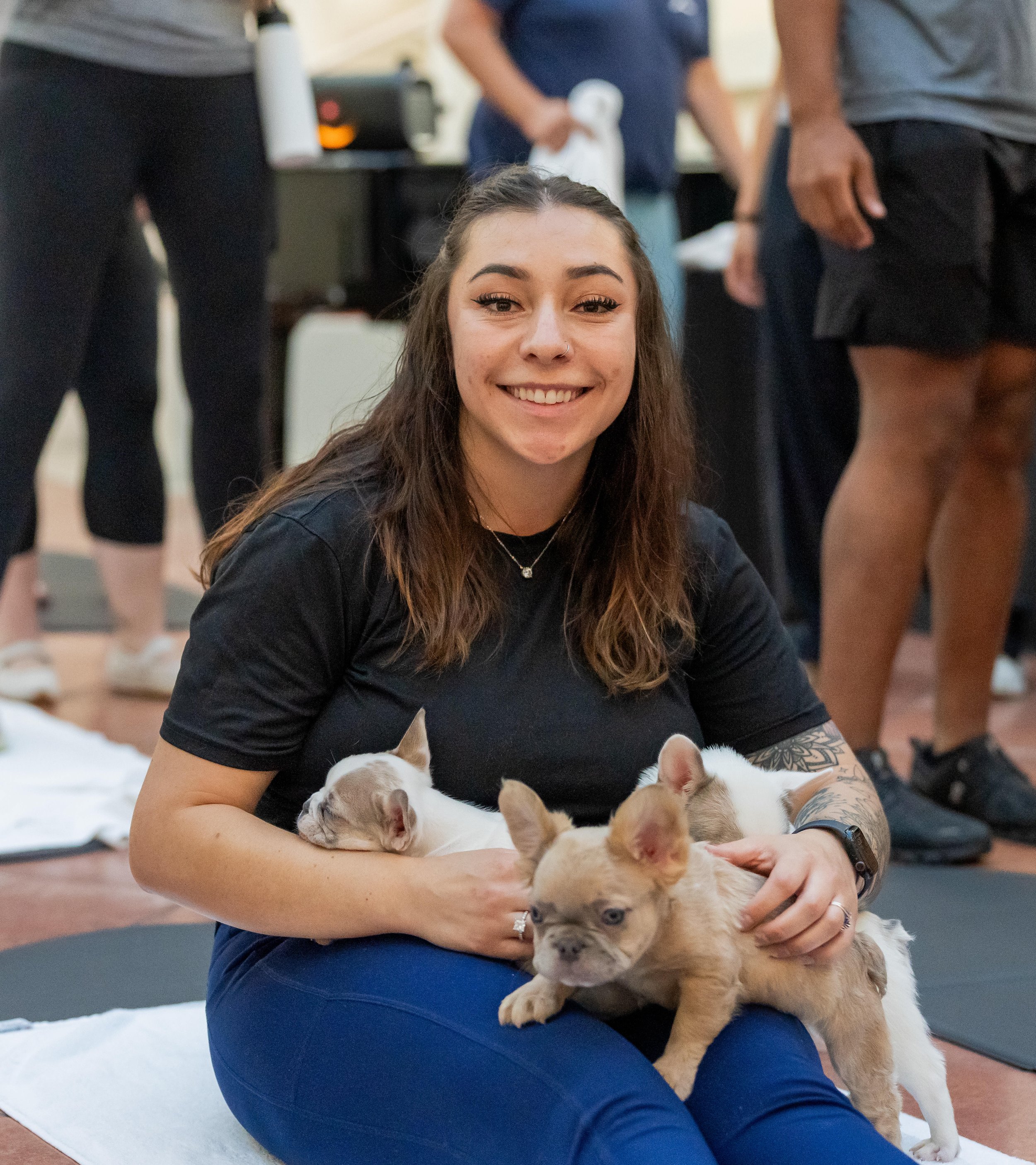 A young woman with long brown hair, smiling, sitting on the floor, surrounded by puppies of different breeds, including a fawn-colored one in her lap and others around her. She is in a room with a few people in the background.