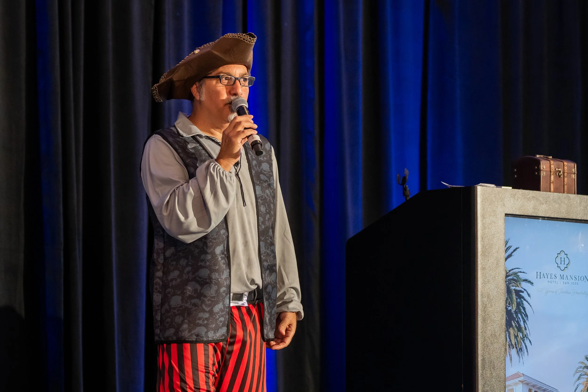 A man dressed in pirate costume standing behind a podium, holding a microphone and speaking, with a dark curtain background and a screen displaying a colorful logo.