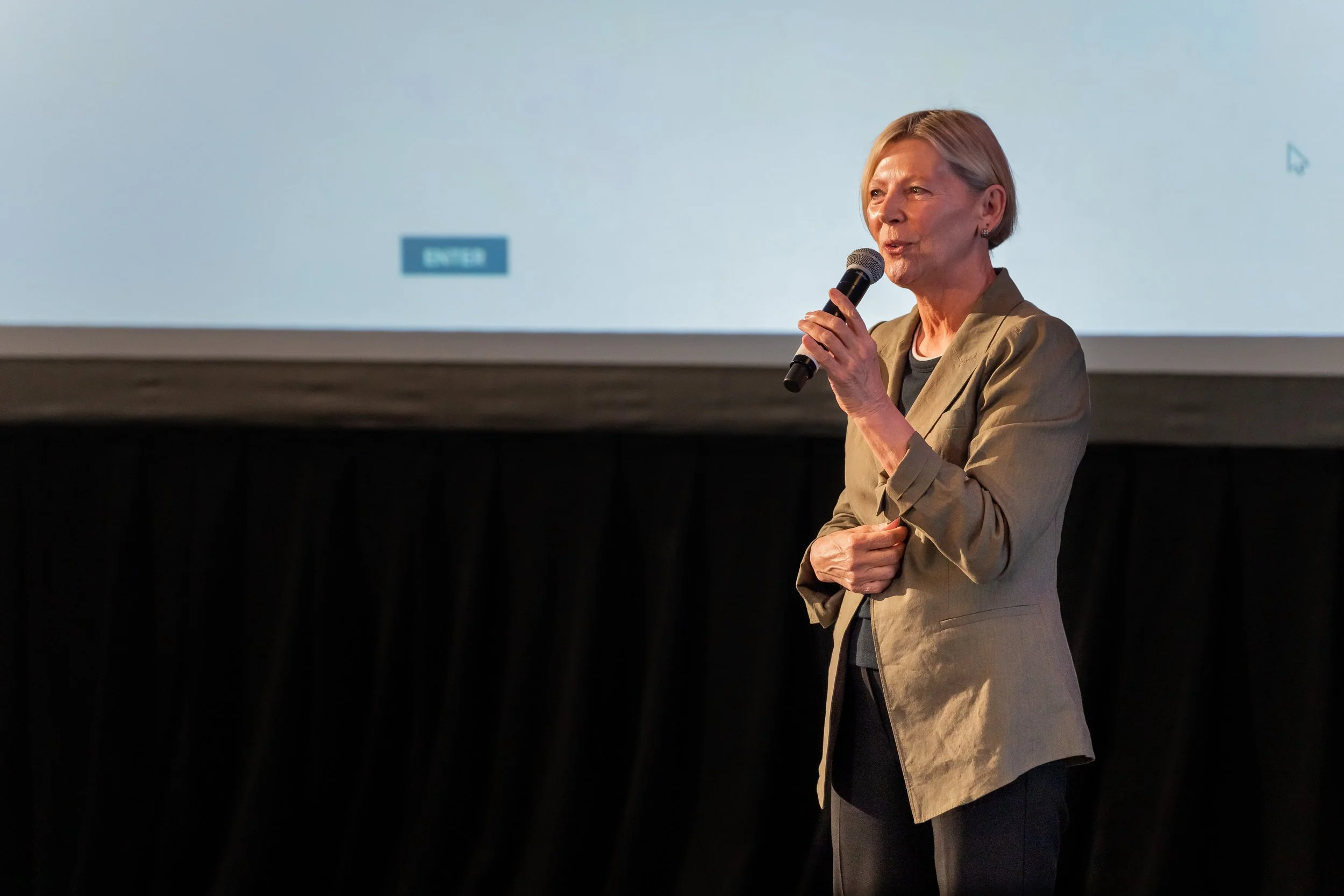 A woman with short blonde hair speaking into a microphone on stage, wearing a beige blazer over a dark shirt, with a large screen behind her.