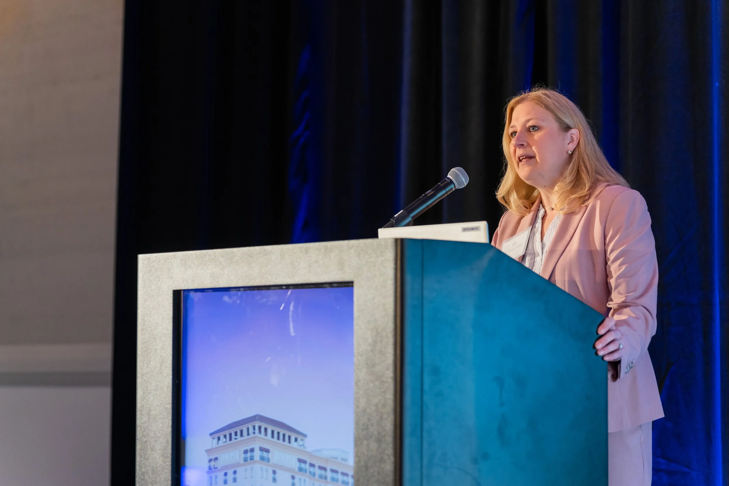 A woman dressed in a pink blazer and white blouse speaking at a conference behind a podium with a microphone. There is a digital screen on the podium showing a building with multiple floors and white columns.