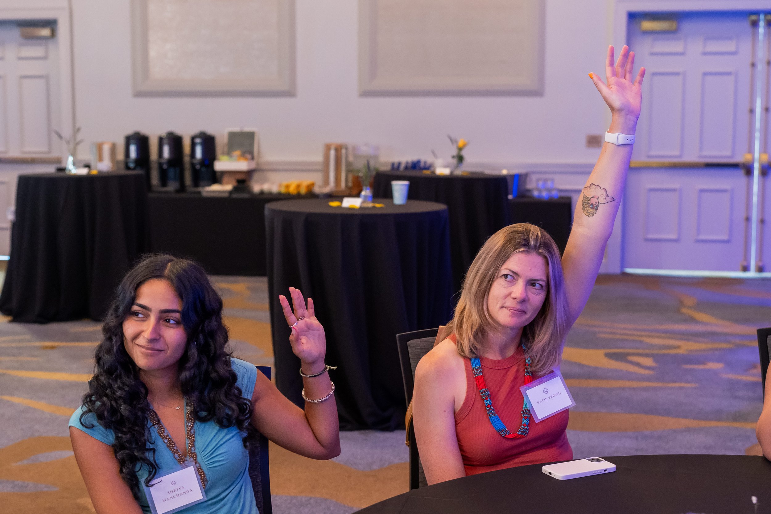 Two women sitting at a table in a conference room, raising their hands. One woman has long, wavy dark hair wearing a blue dress, and the other has straight blonde hair wearing a red dress with a name tag. Background includes a buffet table with coffe