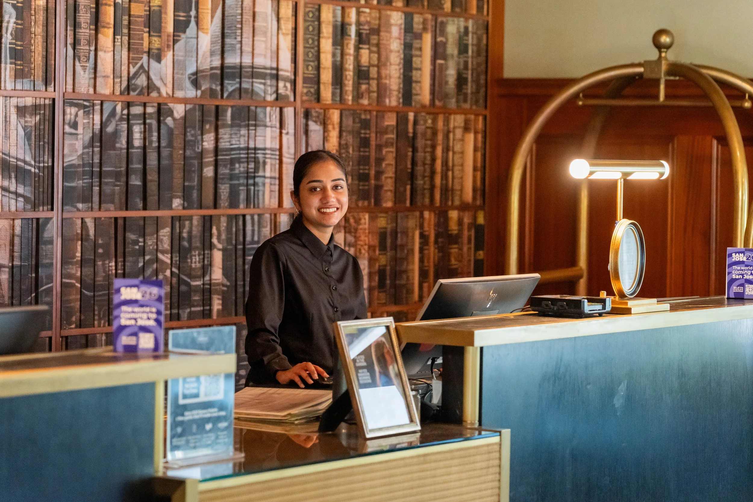 A woman with dark hair in a bun, wearing a black shirt, standing behind a reception desk in a library, smiling at the camera. The background features wooden bookshelves filled with numerous old books and a decorative, vintage-style round mirror with 