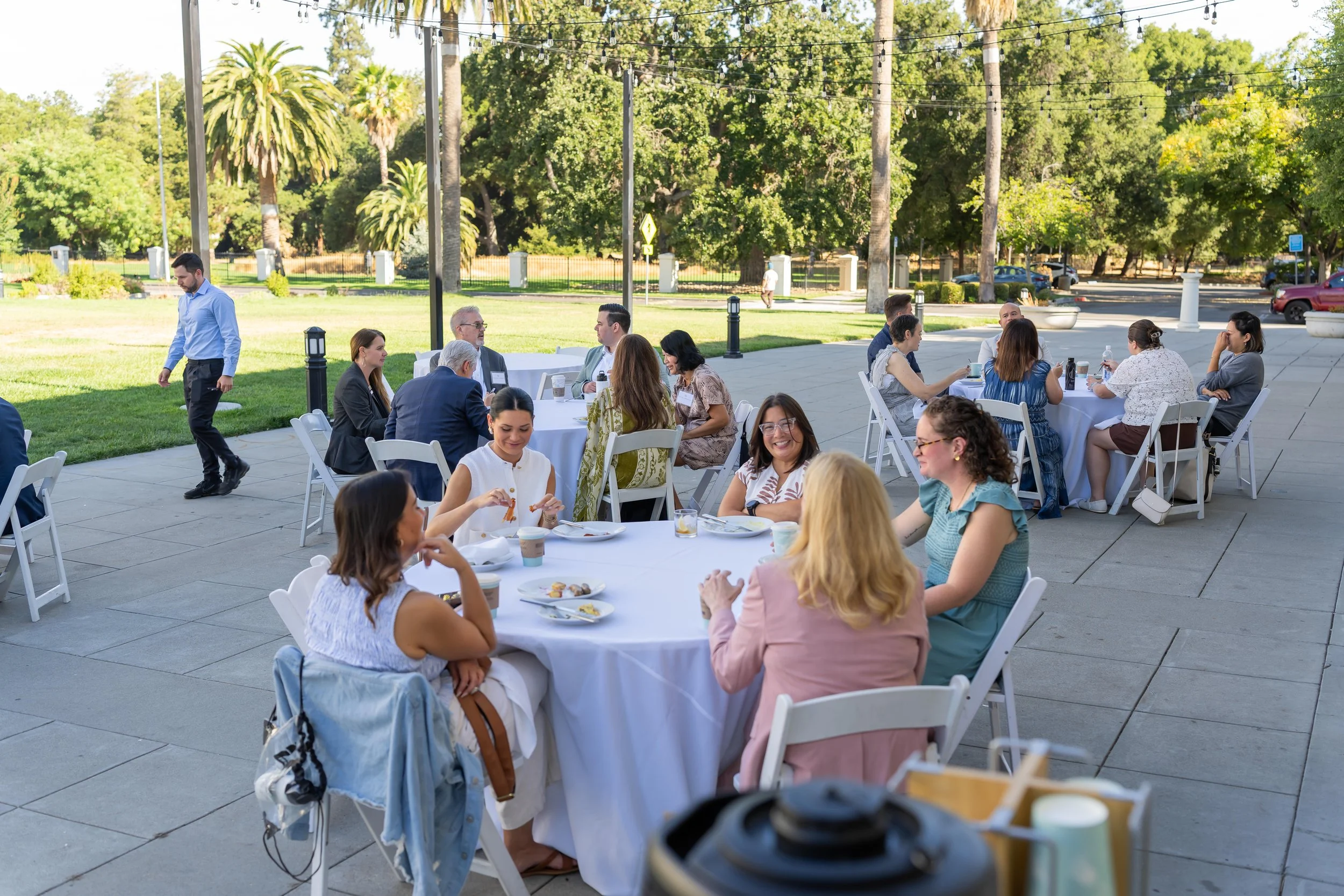 People sitting at white round tables outdoors, enjoying a meal at a garden party or event with string lights overhead and lush green trees in the background.