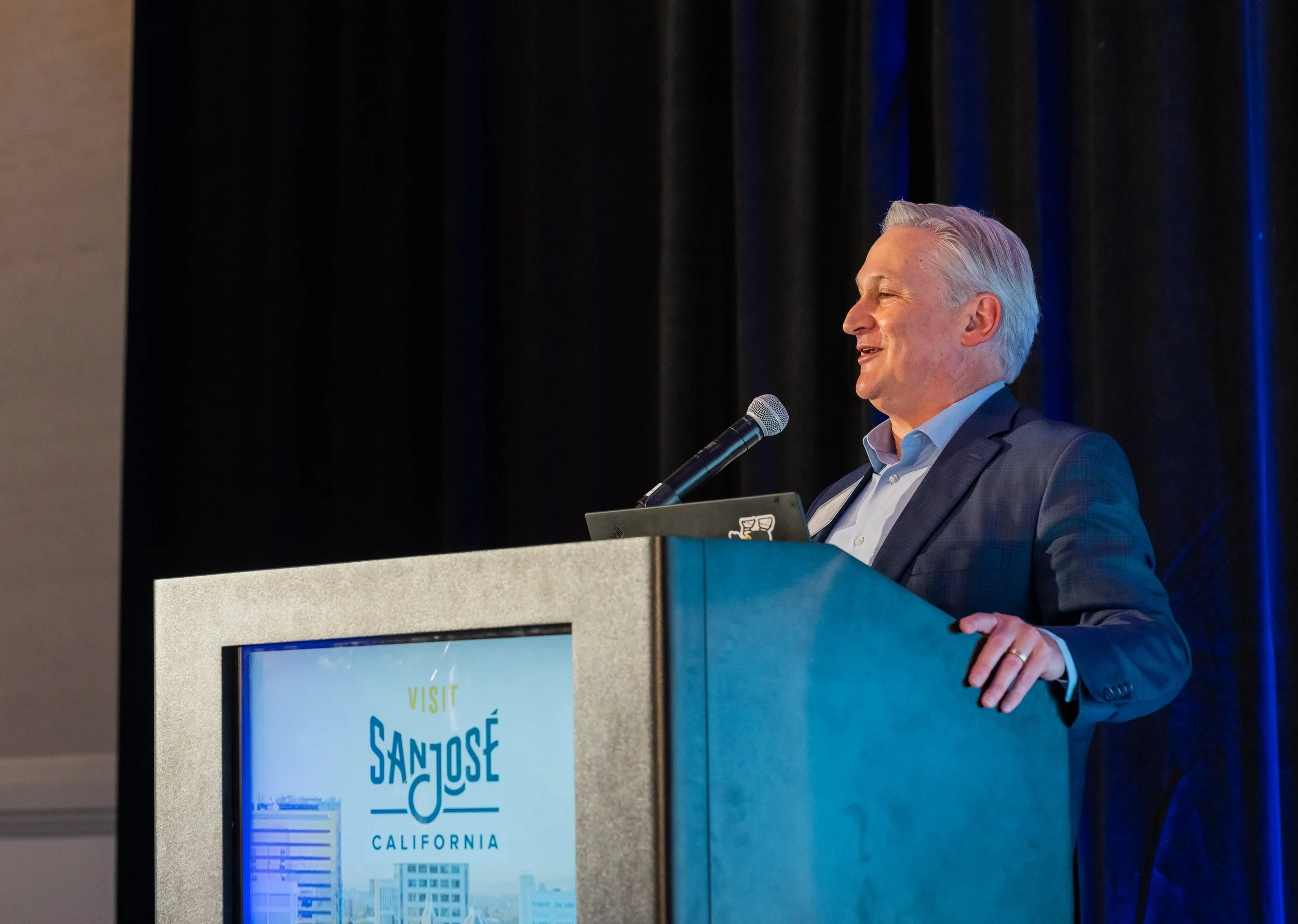 A man with gray hair in a blue suit and white shirt standing at a podium with a microphone, smiling while giving a speech. Behind him is a black curtain, and a blue display screen with the text 'Visit San José California' and an illustration of city 