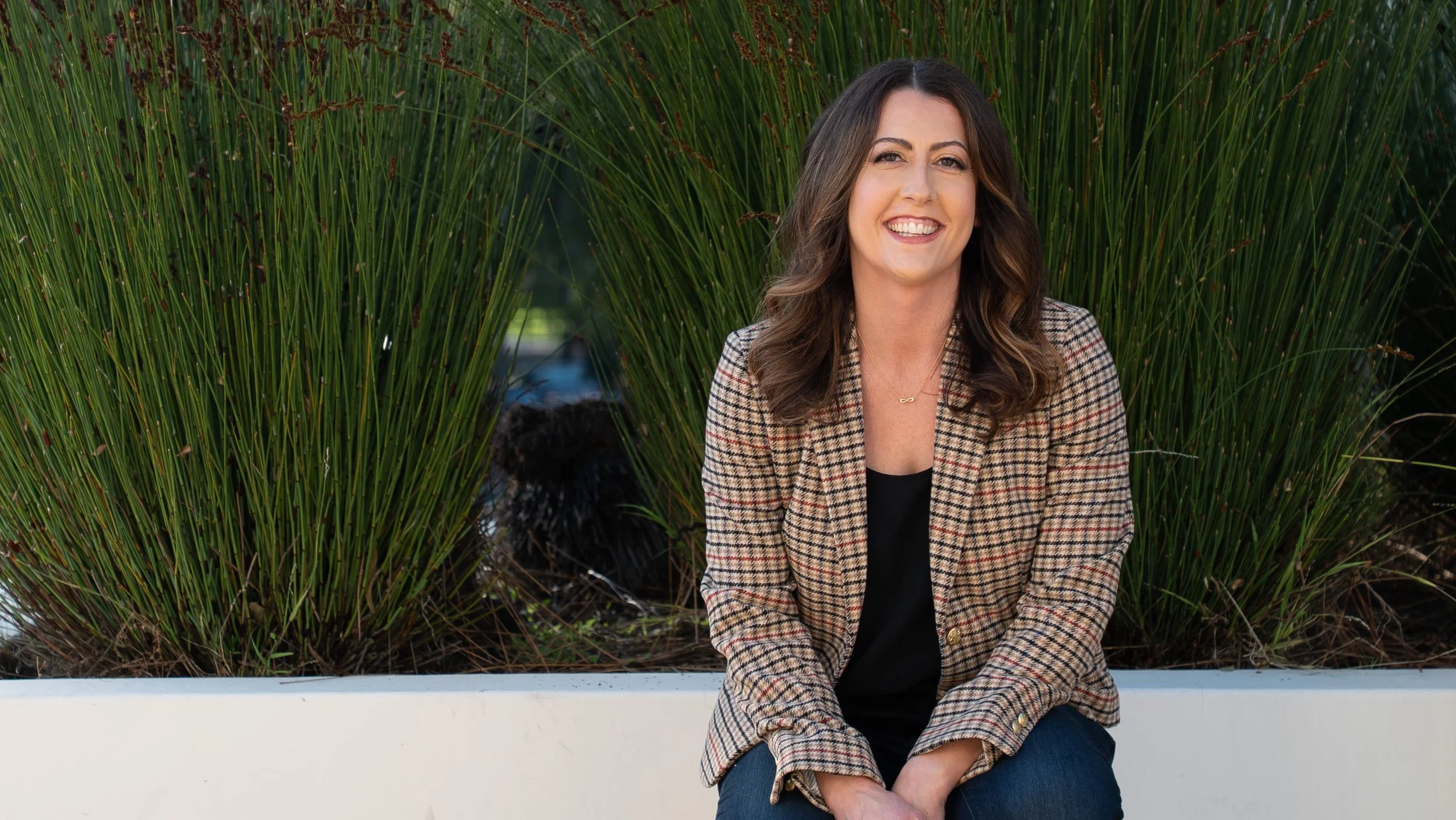 A smiling woman with shoulder-length wavy brown hair, wearing a plaid blazer and black top, sitting in front of tall green grass plants.