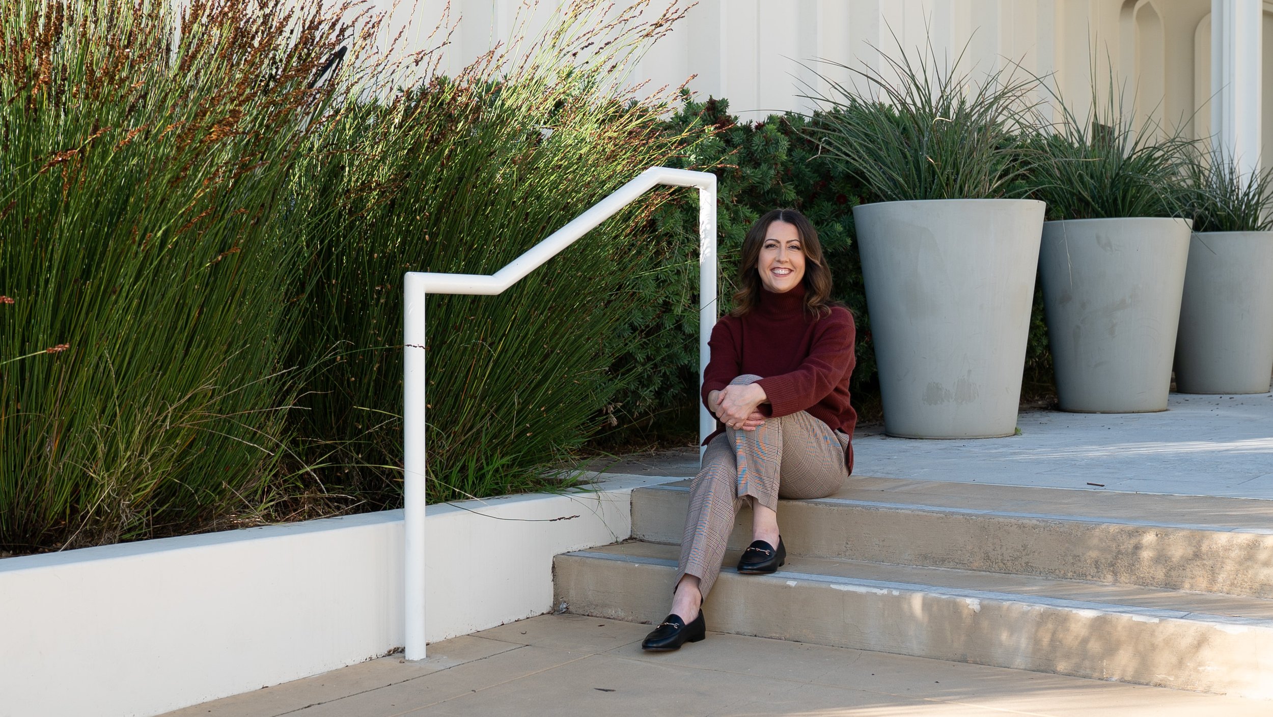 A woman sitting on outdoor steps next to large planters and tall green plants, smiling at the camera.