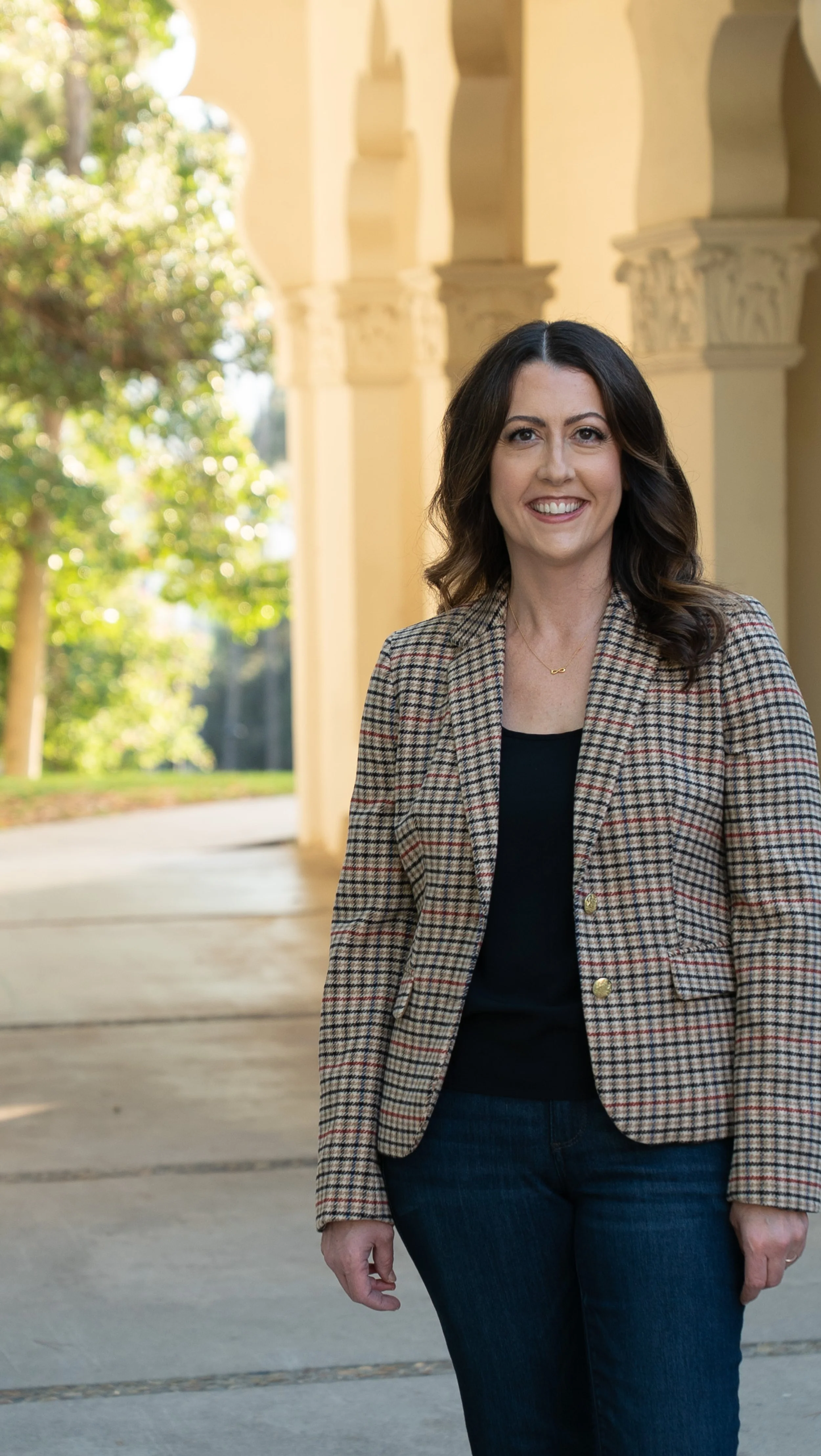 A woman with dark brown hair smiling outdoors with yellow building and green trees in the background.