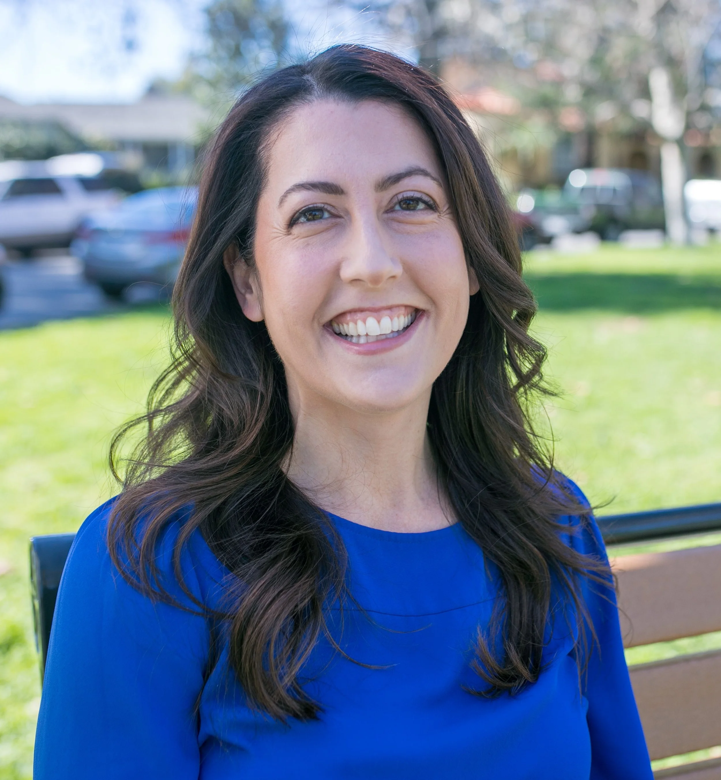 A woman with dark, wavy hair smiling at the camera, wearing a blue top, sitting on a park bench with a grassy area and parked cars in the background.