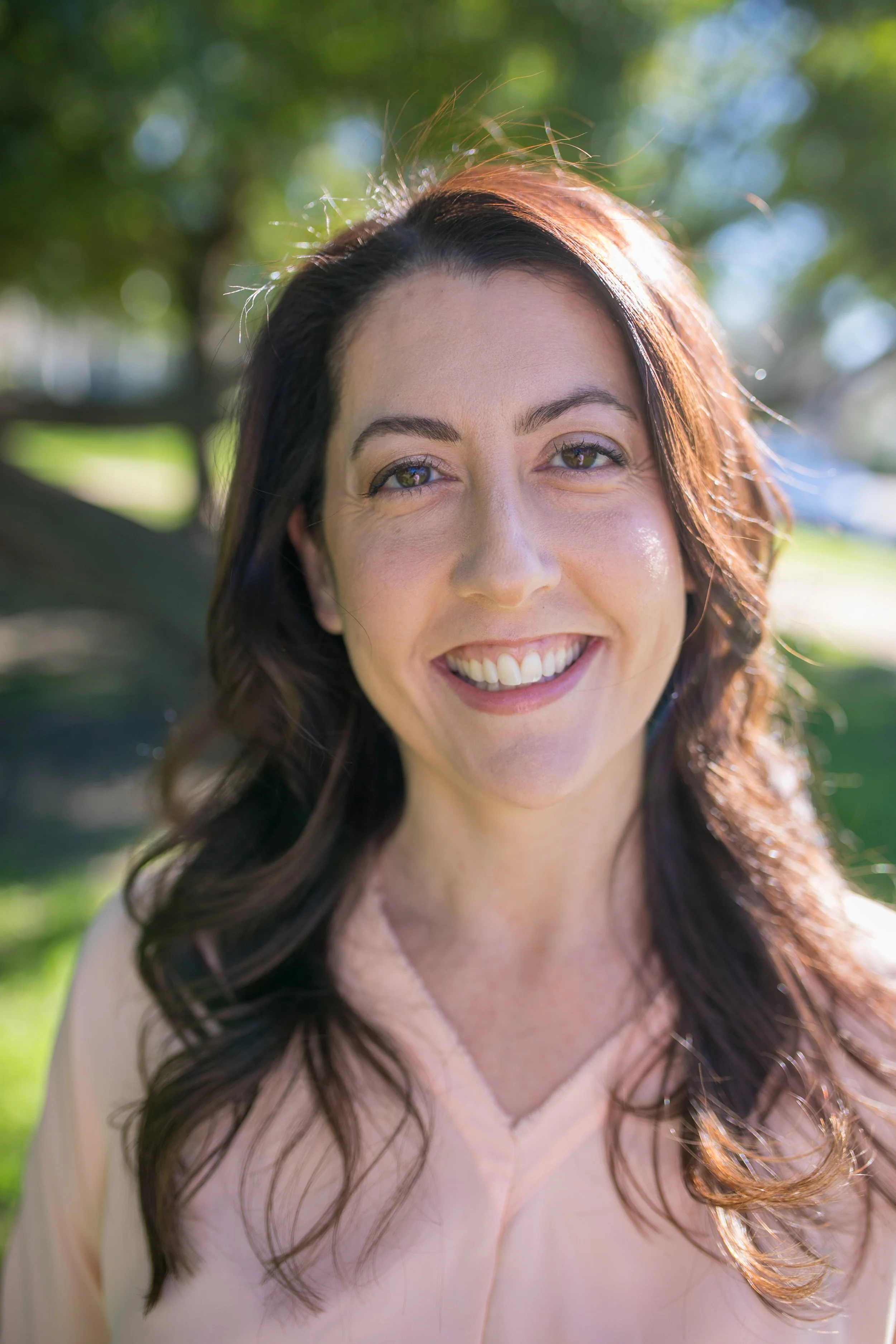 A woman with long brown hair smiling outdoors with green trees and sunlight in the background.