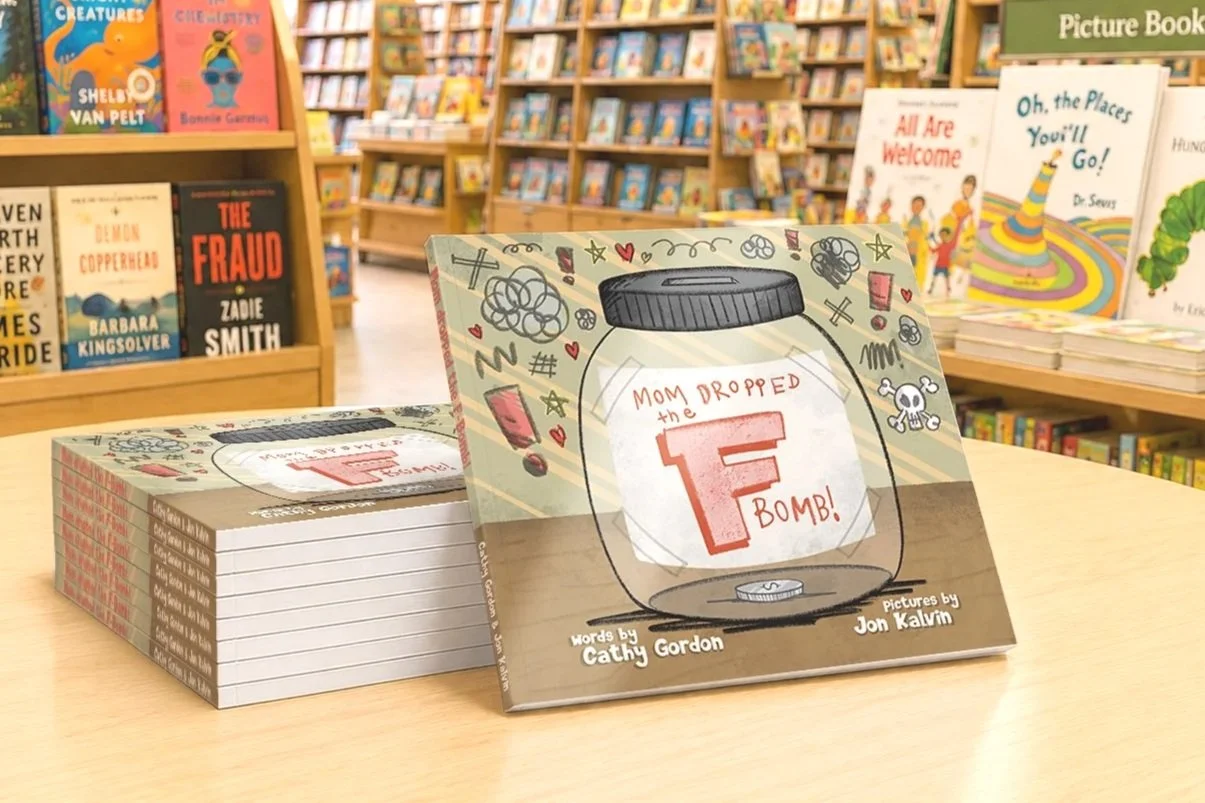 Children's picture book titled "Mom Dropped the F Bomb!" with illustrations of a glass jar, hearts, stars, and a skull on the cover, displayed on a wooden table in a bookstore aisle surrounded by books.