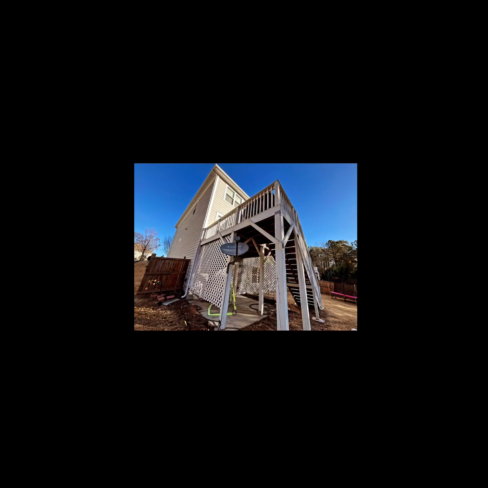 Backyard view of a two-story house with a wooden deck and stairs, and a satellite dish, under a clear blue sky.