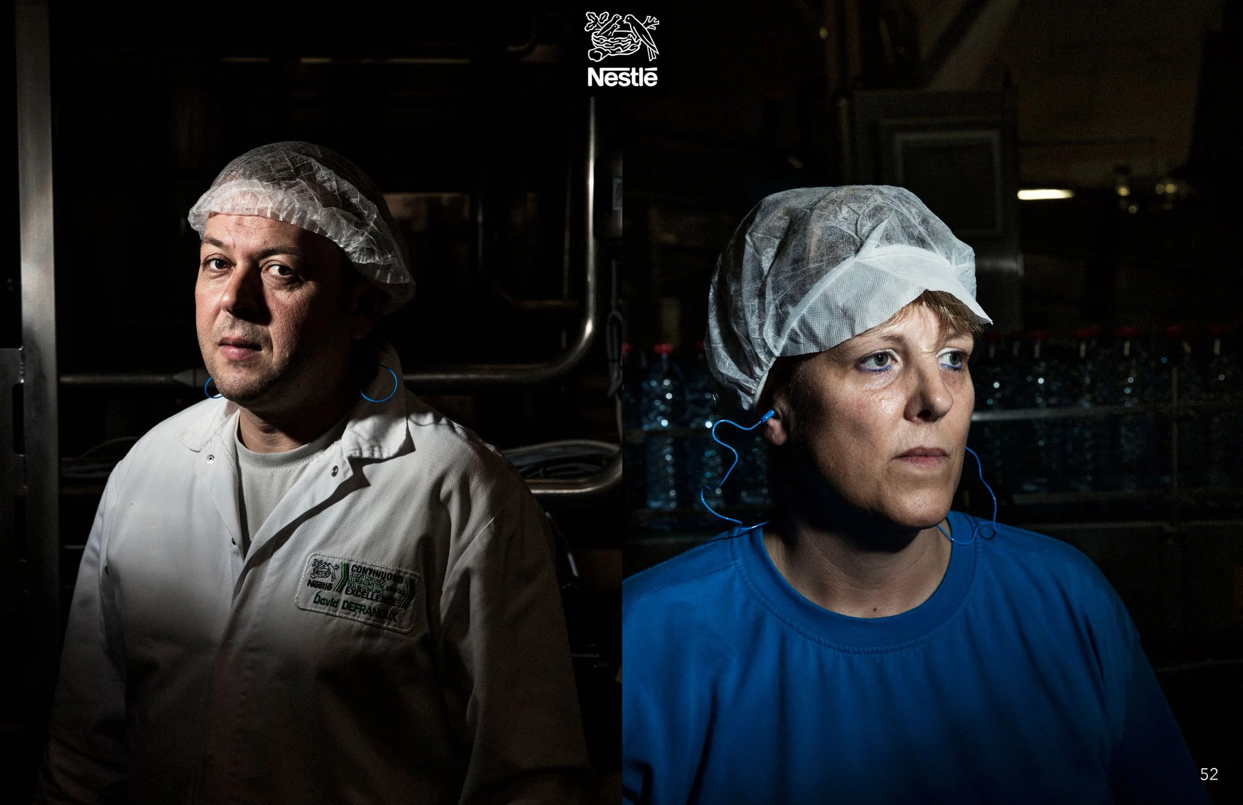 Two people, a man and a woman, wearing hairnets and blue ear protection, standing in a dark industrial kitchen or food production facility, with shelves of bottled water behind them. The man is wearing a white chef jacket with a name tag, and the wom