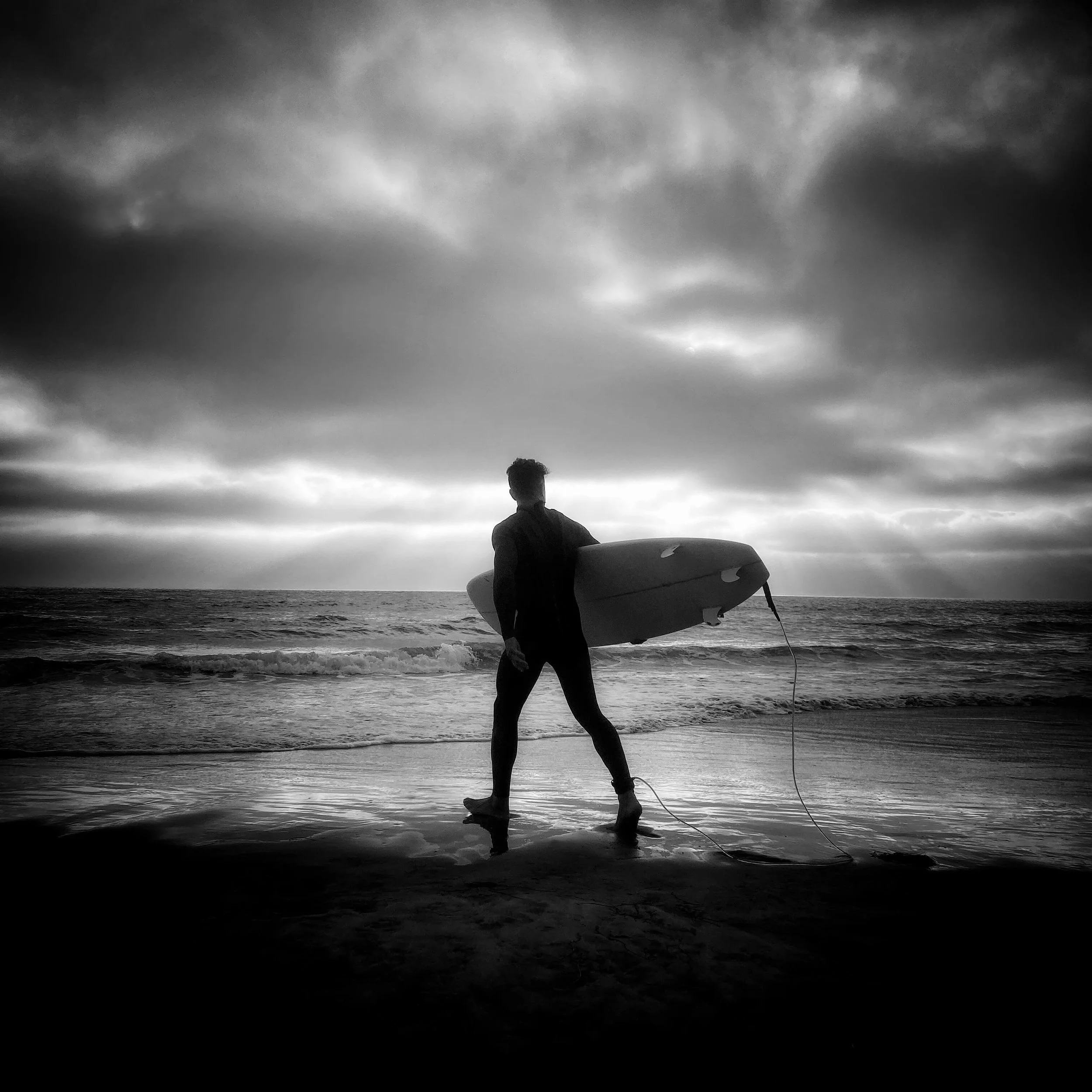 A person walking on the beach with a surfboard, facing the ocean under a cloudy sky.