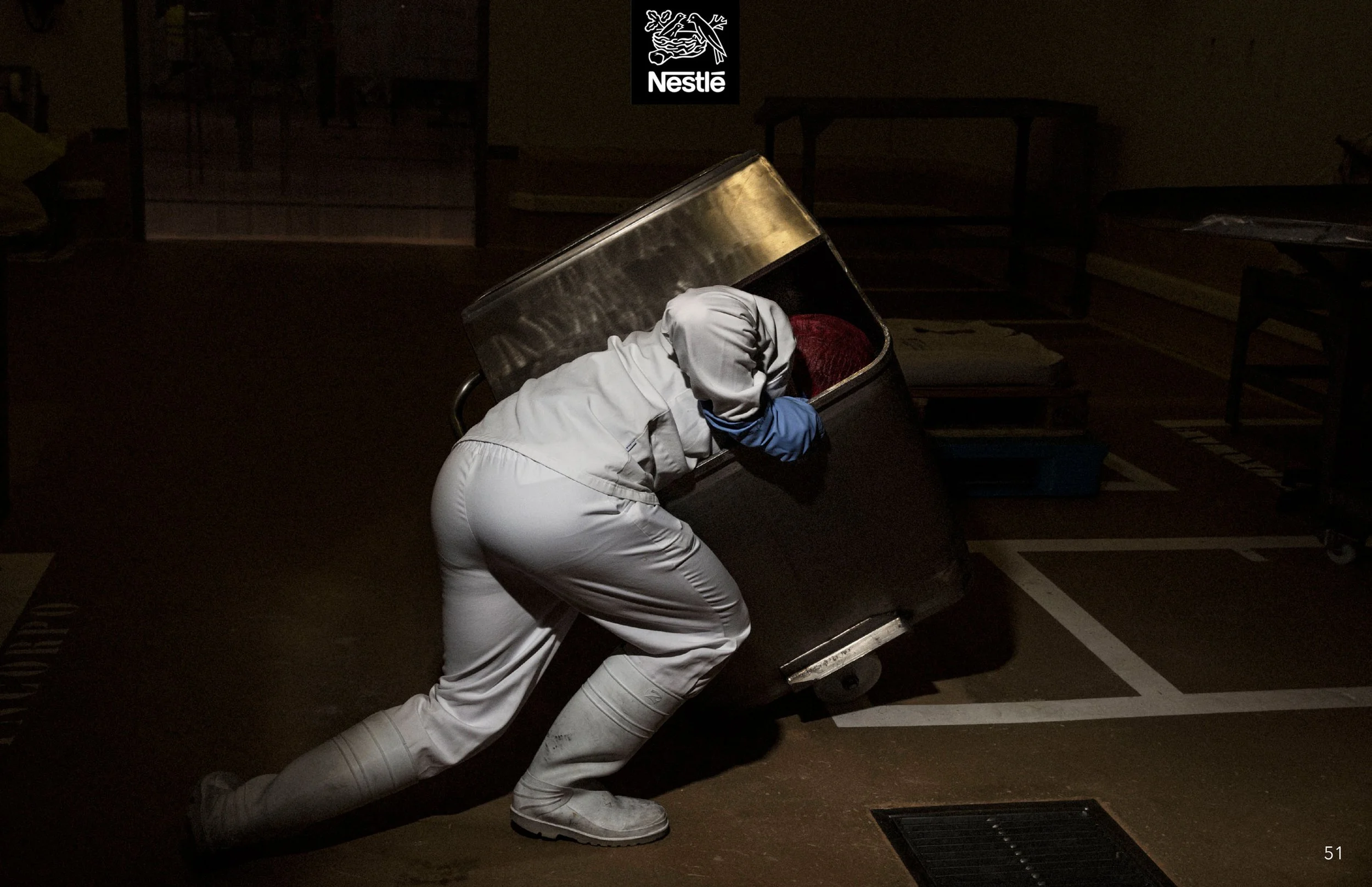 A person in white coveralls and a red beanie pushing a metal cart in a dark room with minimal lighting. The room appears to be a storage or utility space with tables and other items in the background. The Nestlé logo is visible in the top center of t