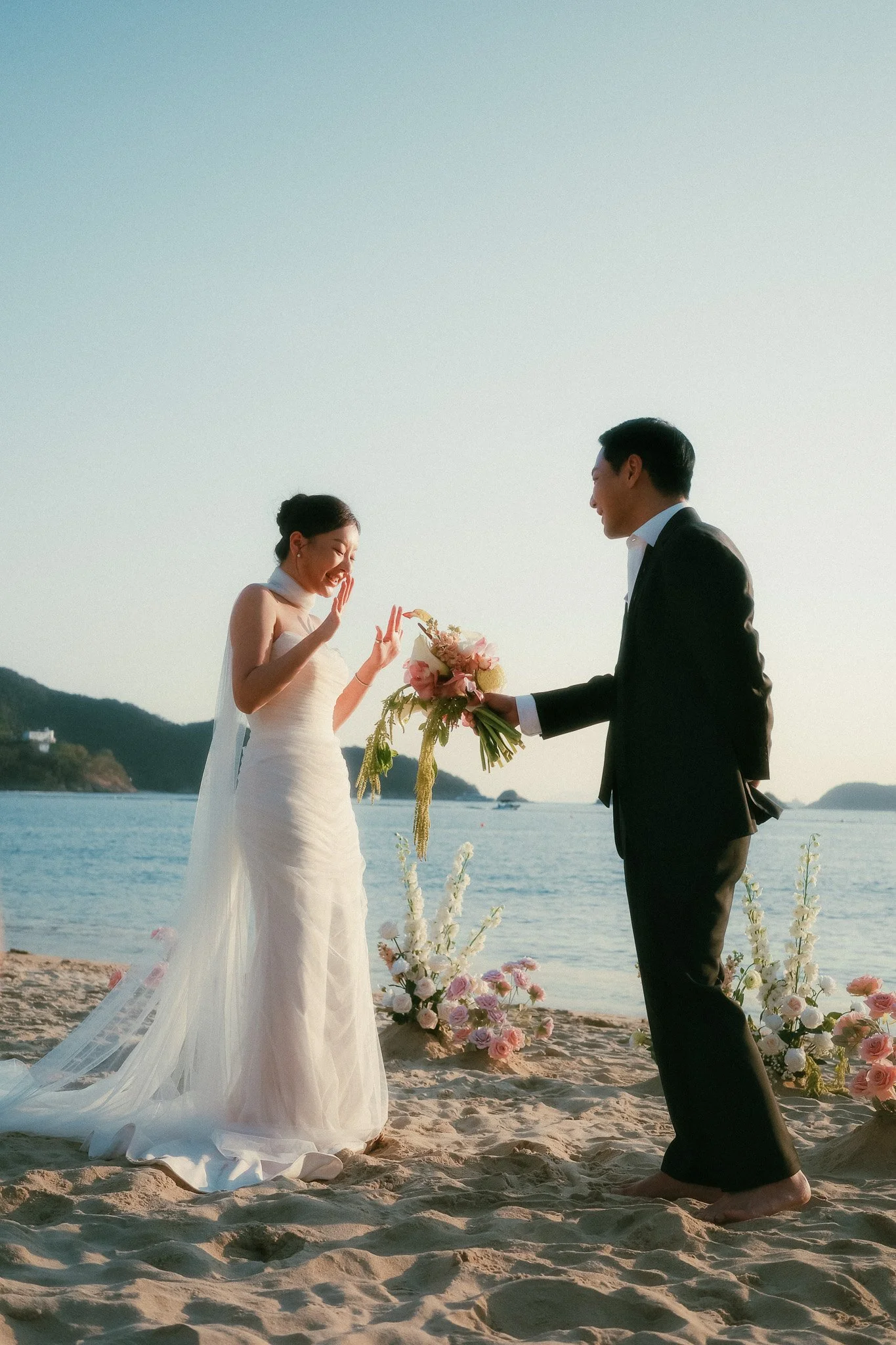 Beach elopement at twilight. One partner handing a bouquet of pink and white flowers to the other, ocean and sand illuminated behind them