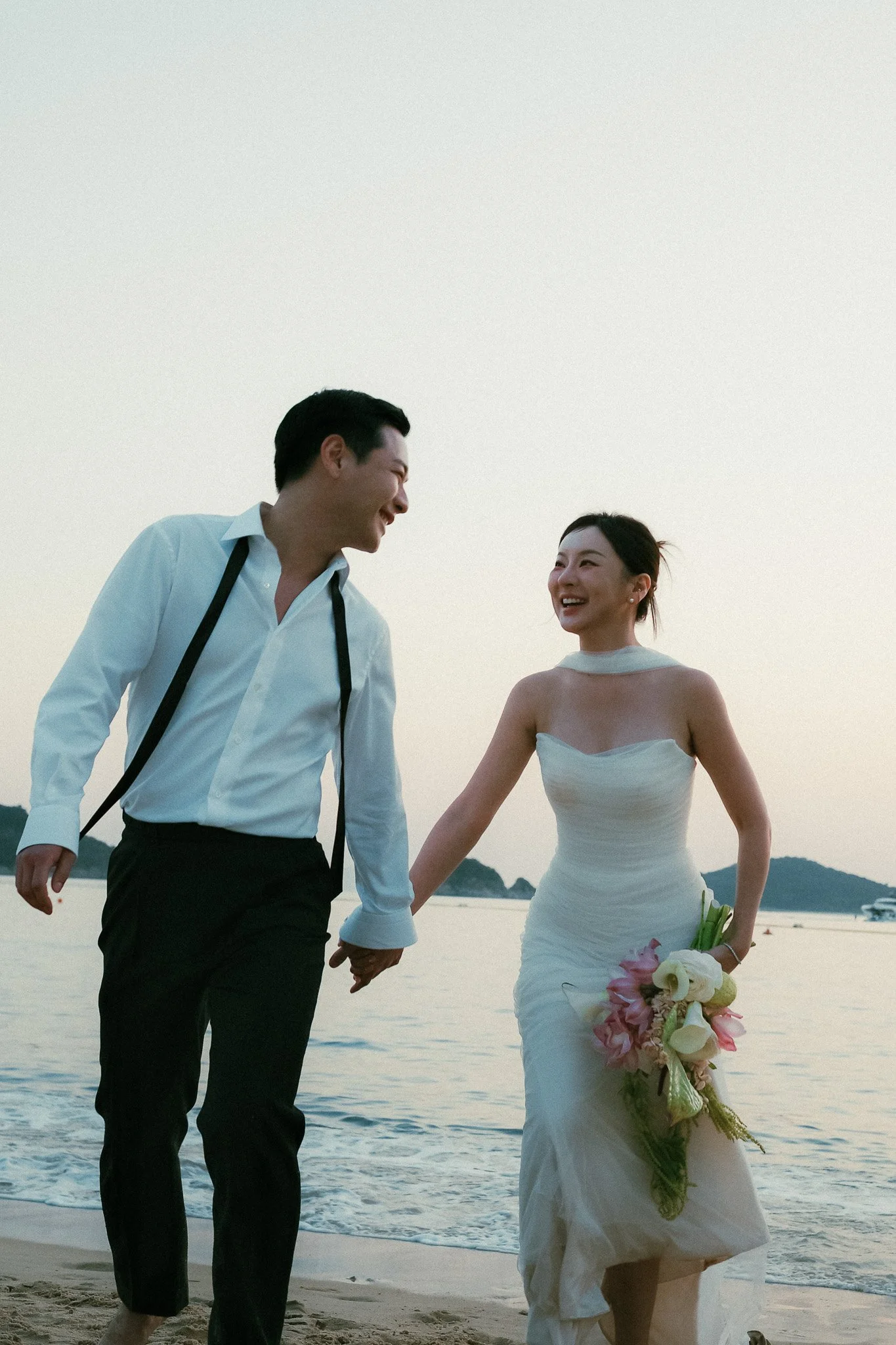 Bridal couple walking hand in hand back toward shore, one partner glancing back at the other, twilight sky behind them