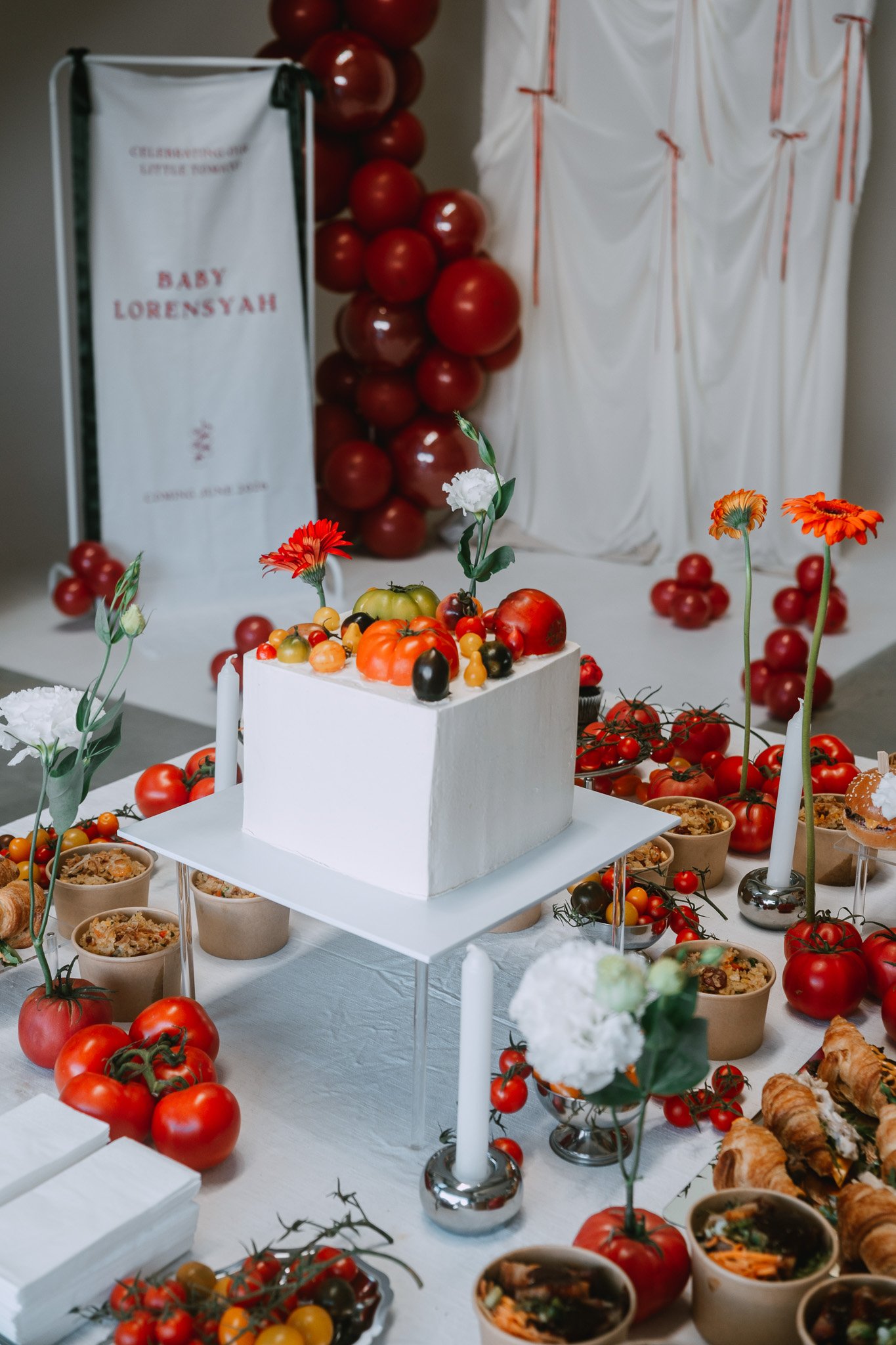 Baby shower table styled in shades of red with white backdrop, red balloon tower visible behind