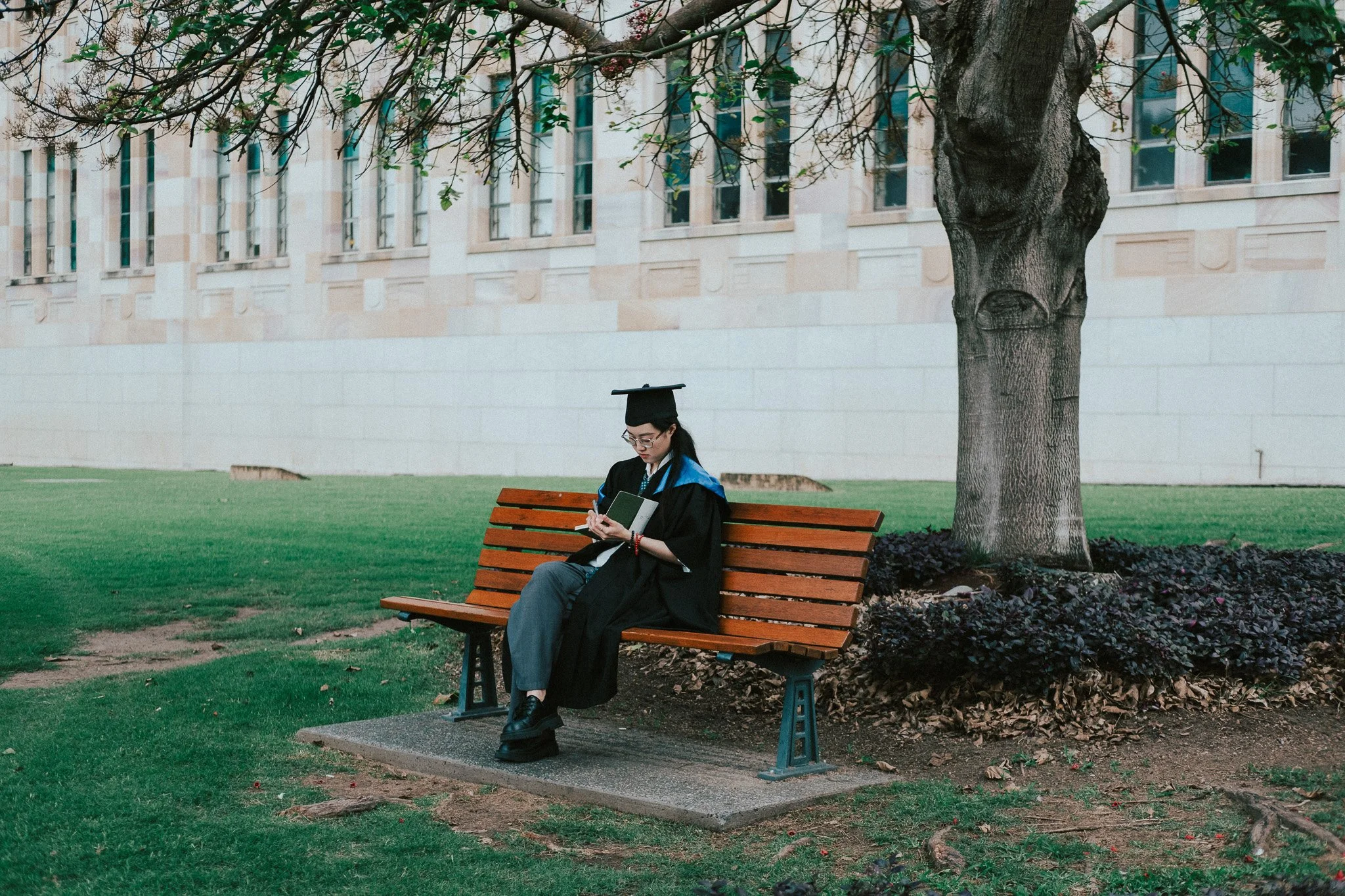 Graduate seated on a wooden bench in graduation robes and mortarboard, grassed grounds of University of Queensland around her