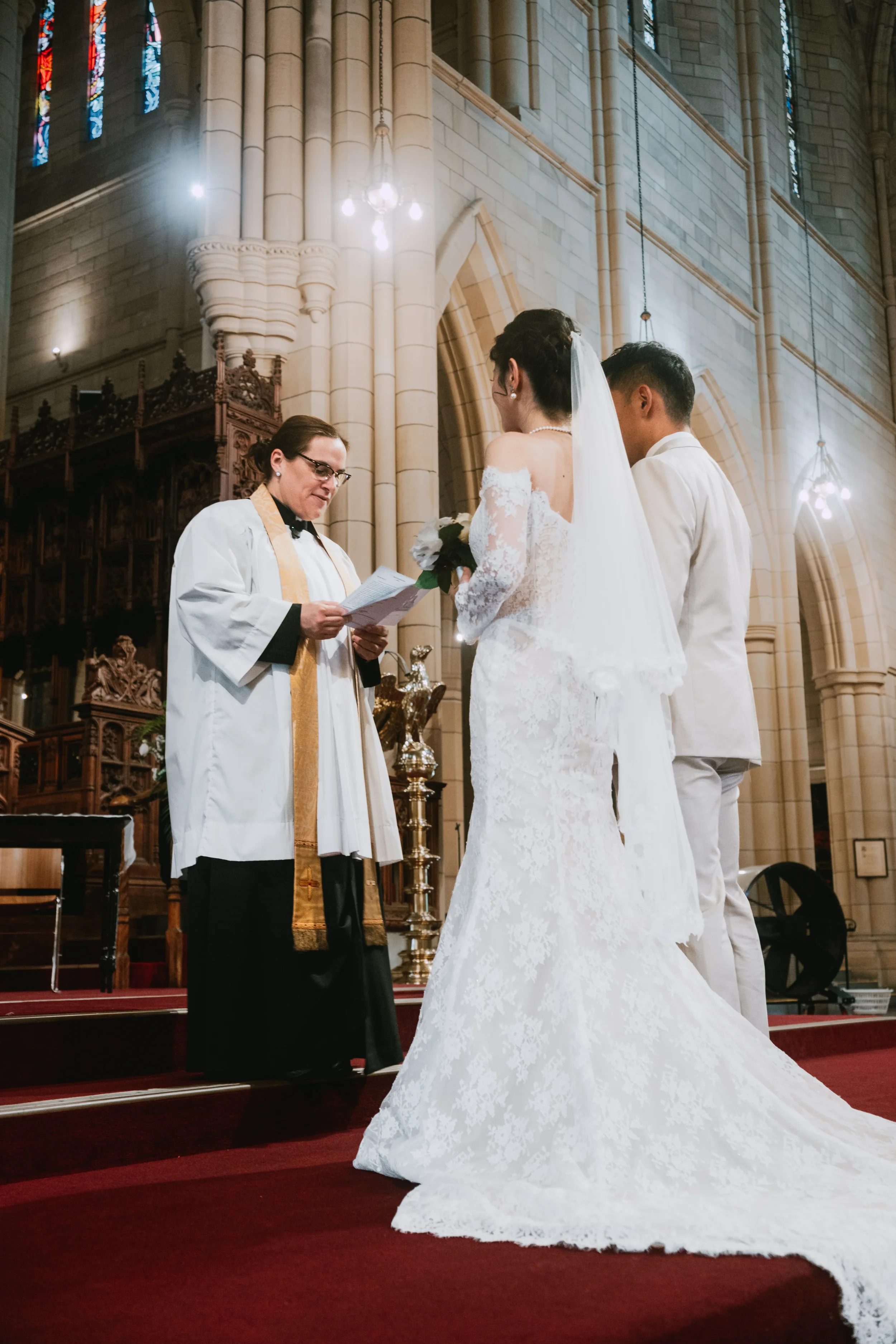 Bridal couple standing before their officiant mid-ceremony, Catholic church interior behind them, chandelier light overhead