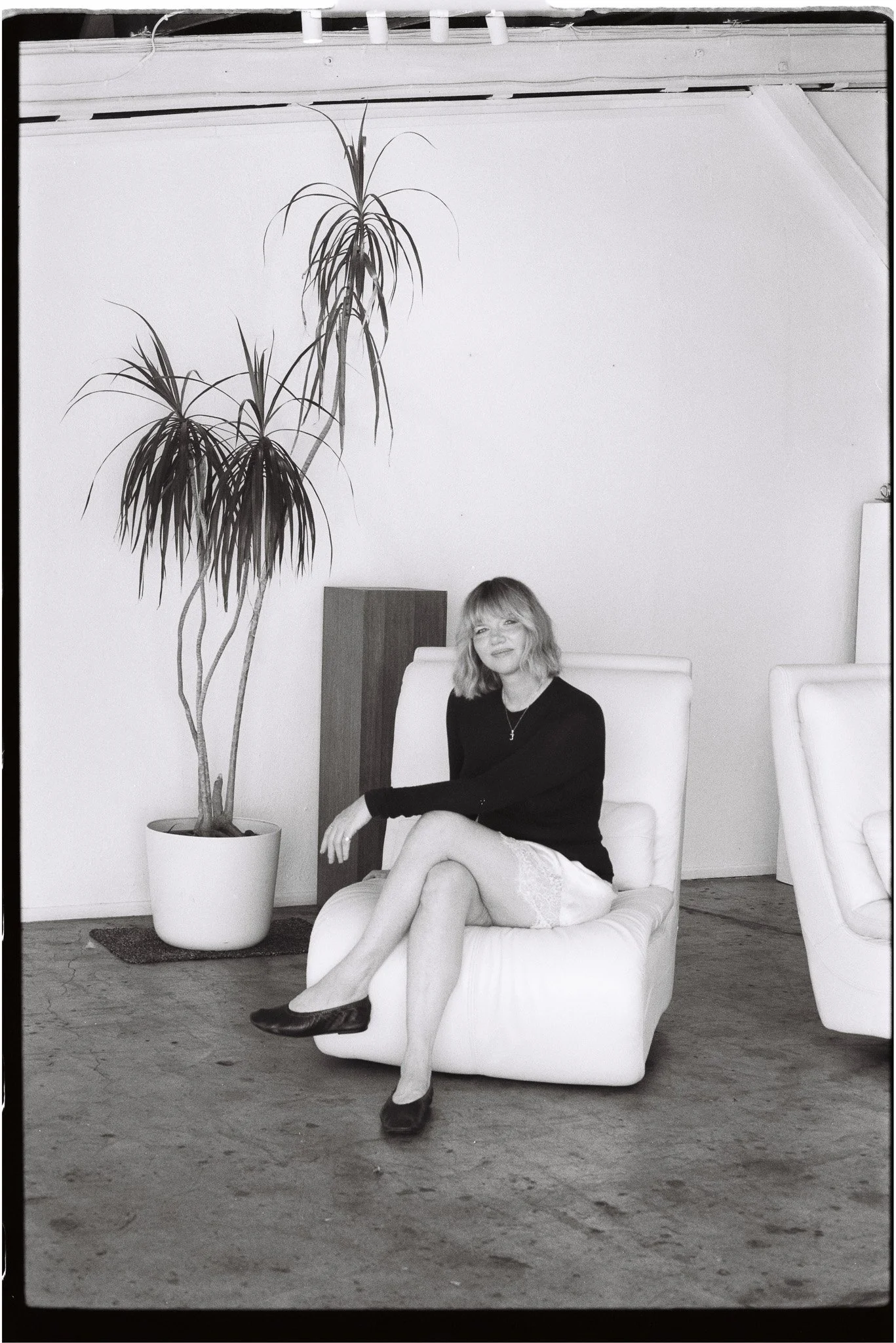 Woman seated cross-legged on a chair, a potted ponytail palm beside her