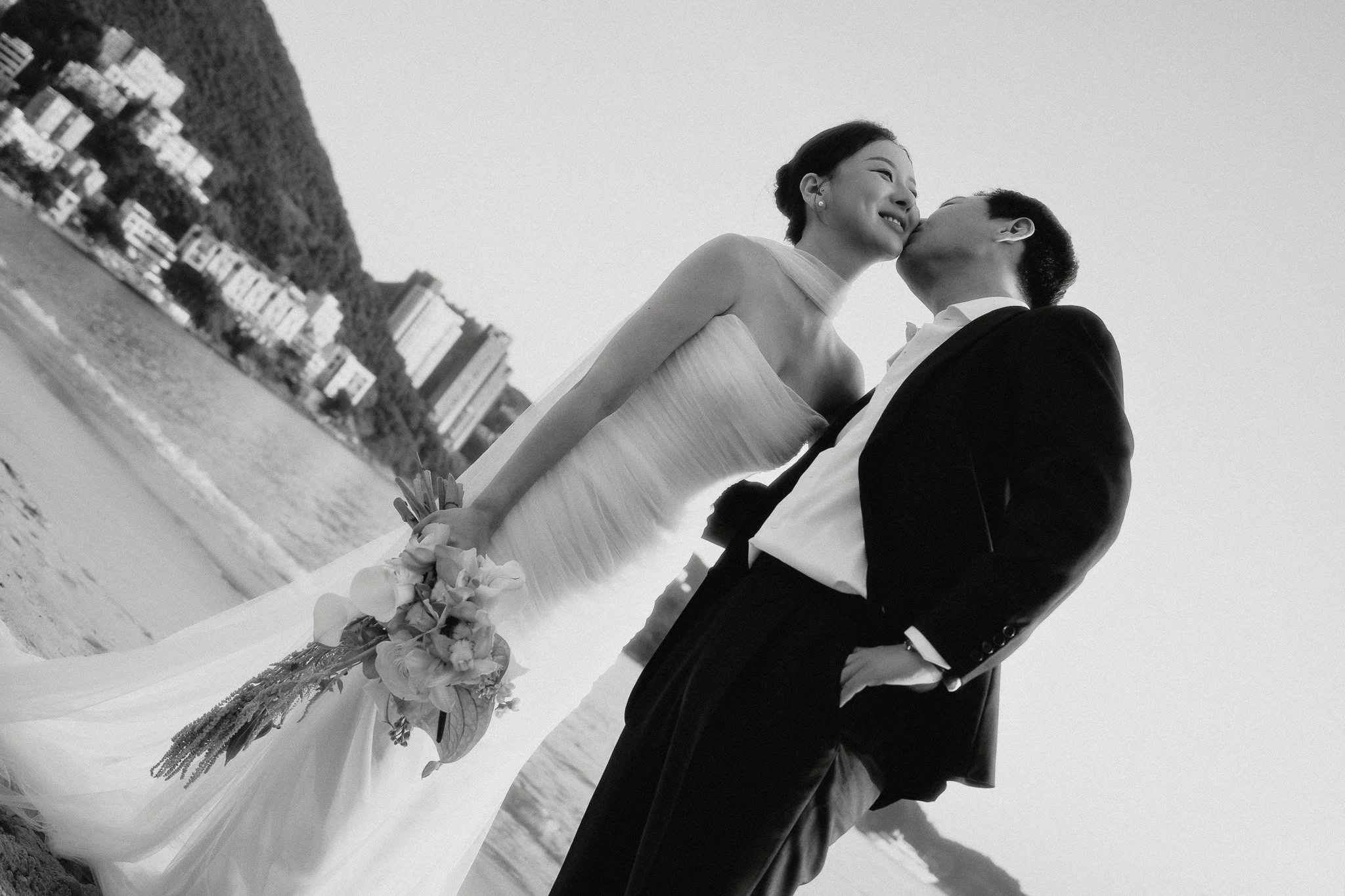 One partner kissing the other on the cheek on the beach, the other holding a bouquet at their side