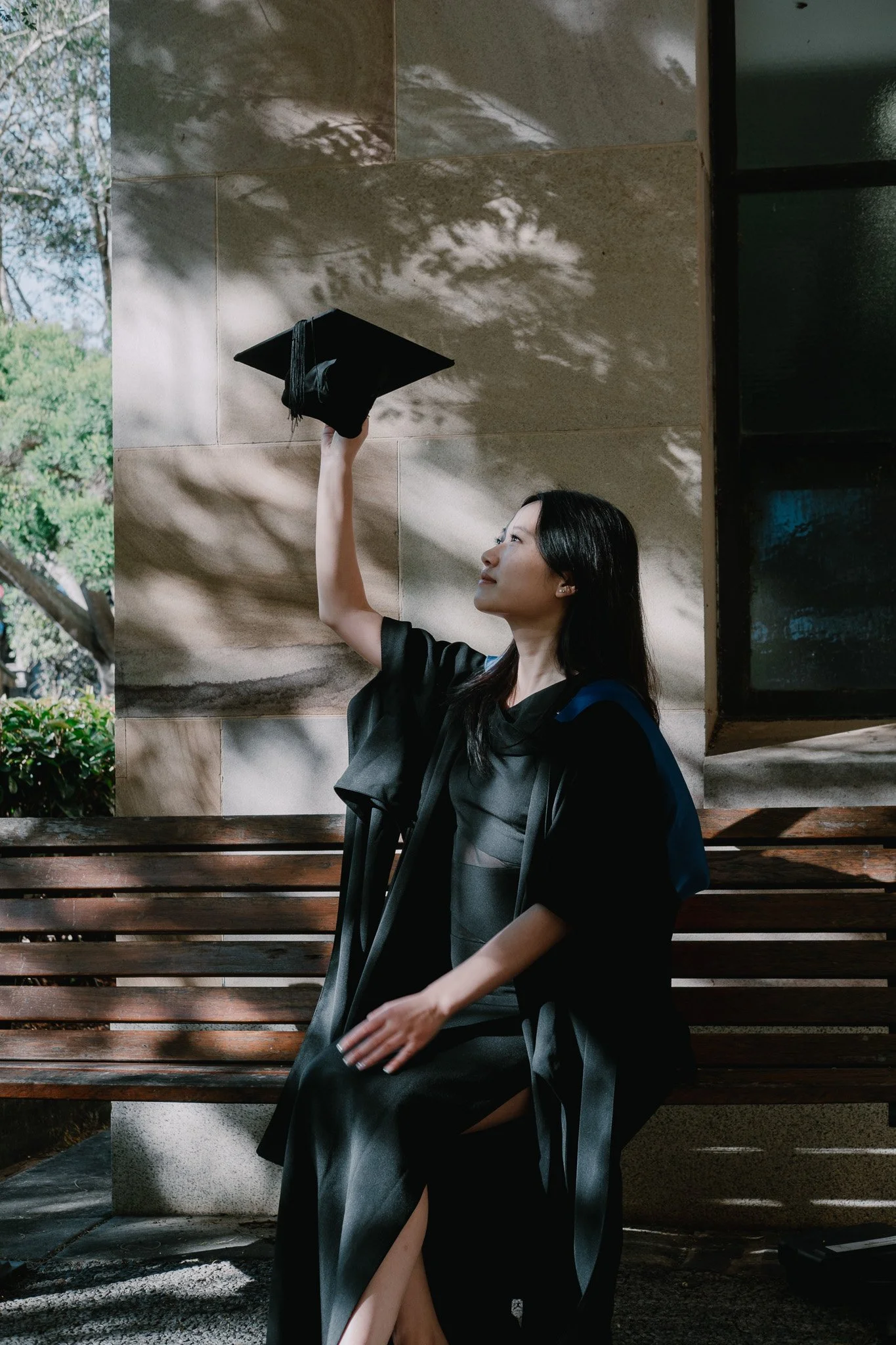 Graduate seated on a bench, holding her mortarboard in one hand, dappled light filtering through the trees above