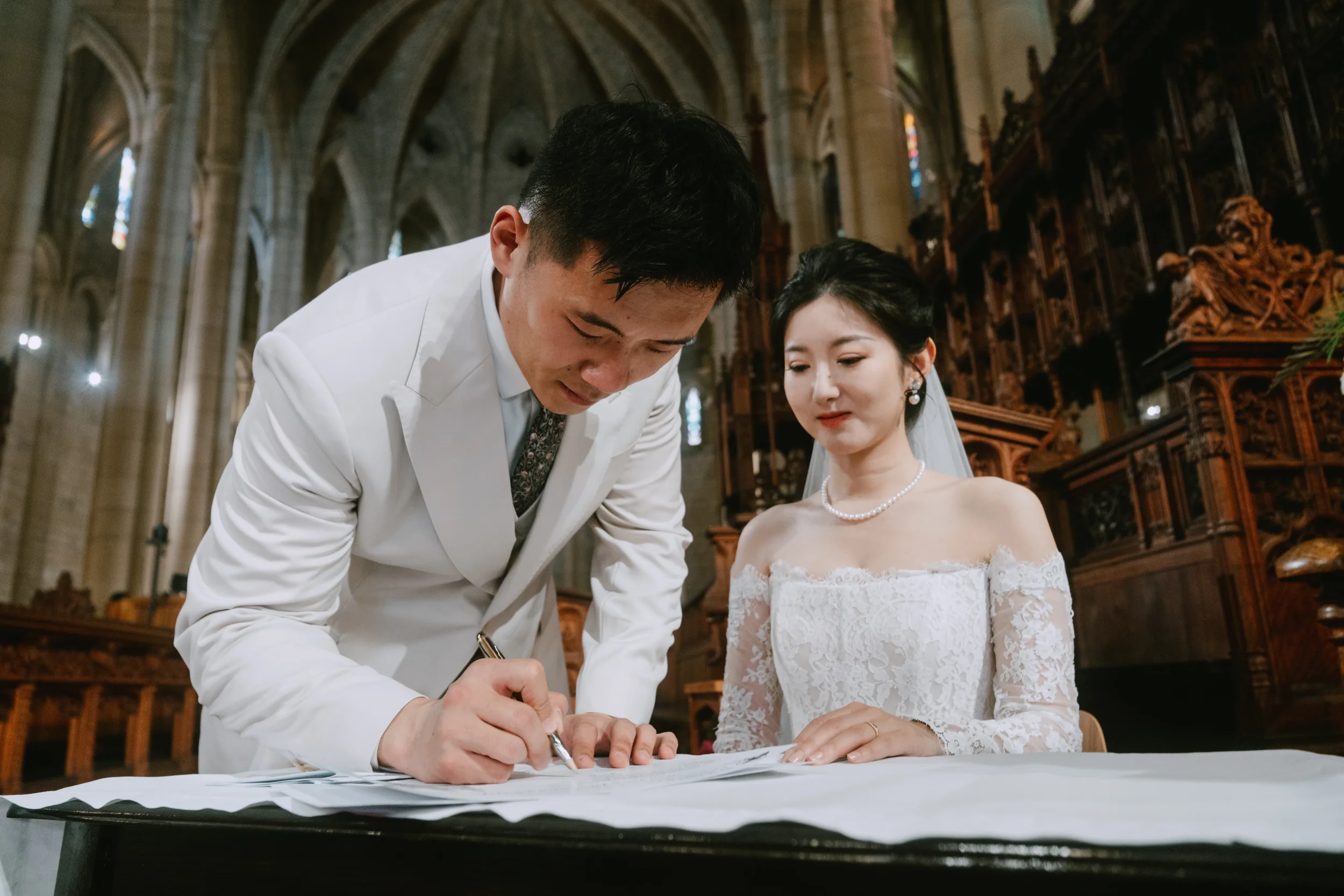Couple signing the marriage certificate, one partner standing and leaning over the document to sign, the other seated beside them