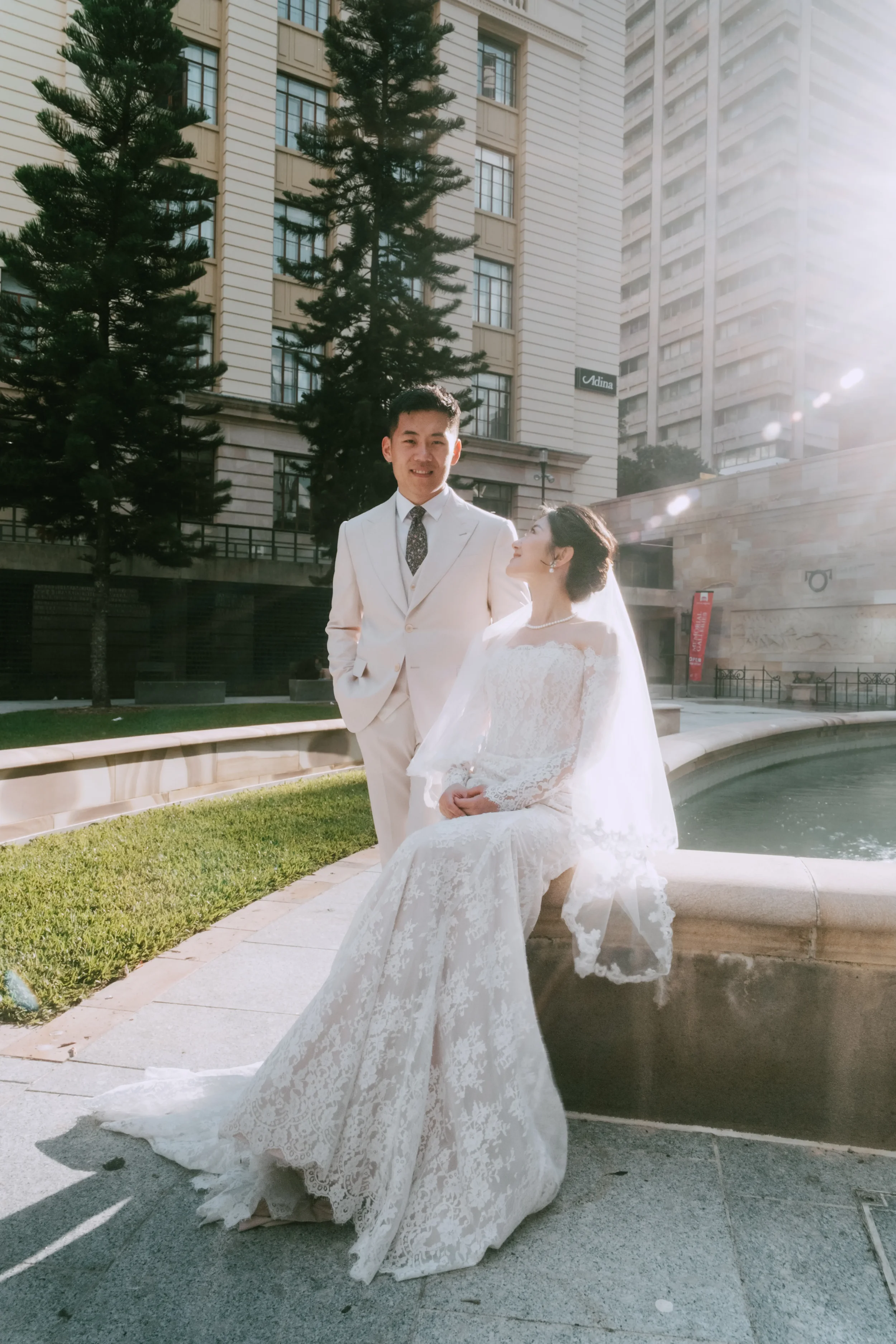 Bridal couple together, one partner seated on a low stone ledge looking up at the other who is standing, both smiling, strong light from the right
