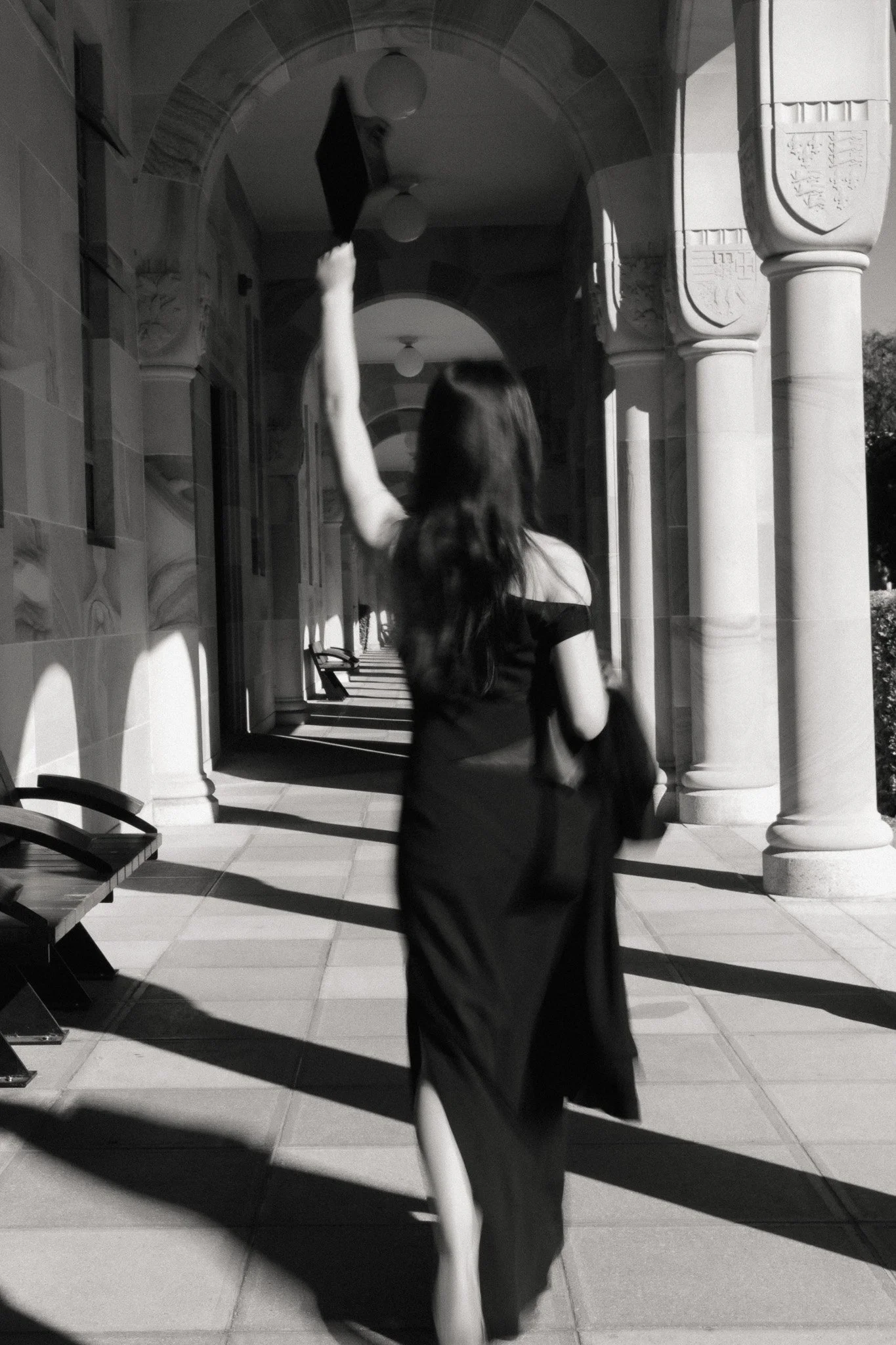 Graduate walking through a covered arcade in University of Queensland, mortarboard raised in one hand, pillar shadows falling across her