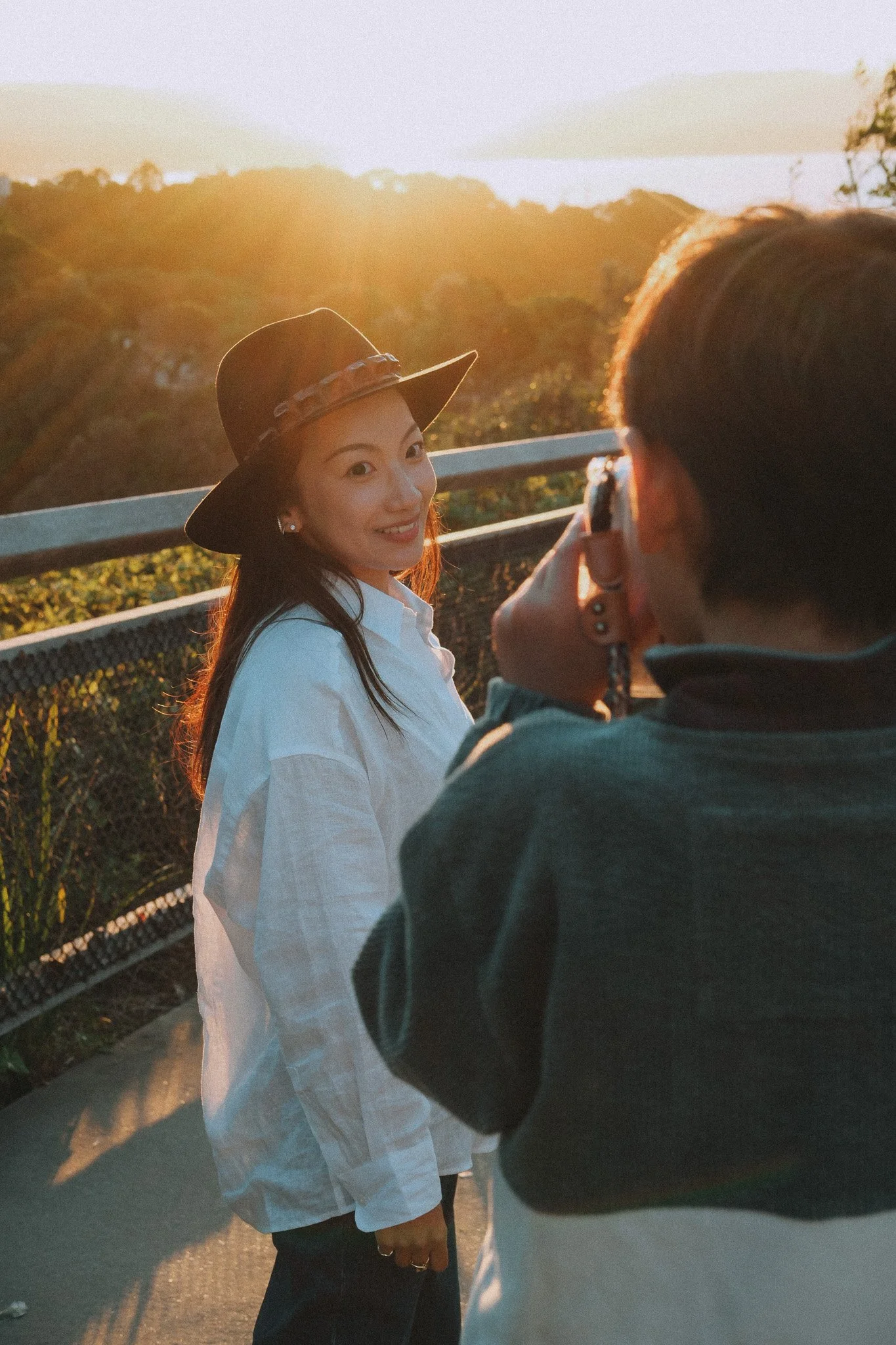 Over the shoulder shot of son, Mum is visible ahead as he photographs her on his Fujifilm Instax, forest and sunset behind her