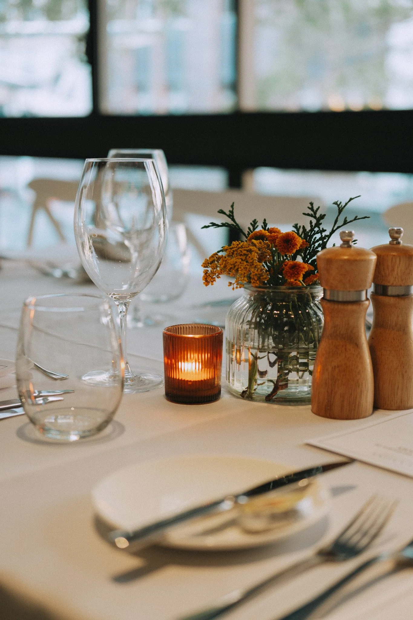 Table set for an engagement party lunch, white plates and silver cutlery, lit orange candle and small orange and yellow floral centrepiece
