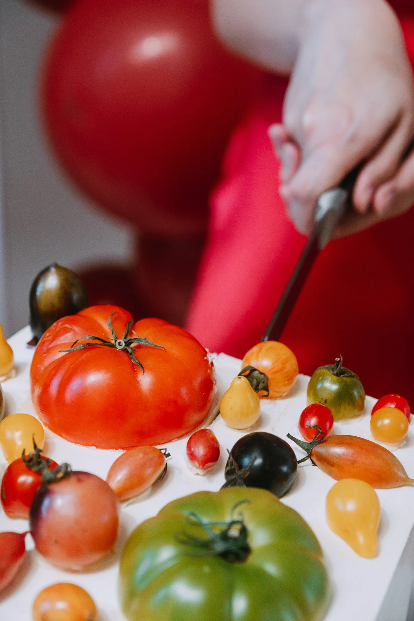 Close detail of hands pressing a knife down to cut a white-iced cake decorated with tomatoes in various colours and sizes