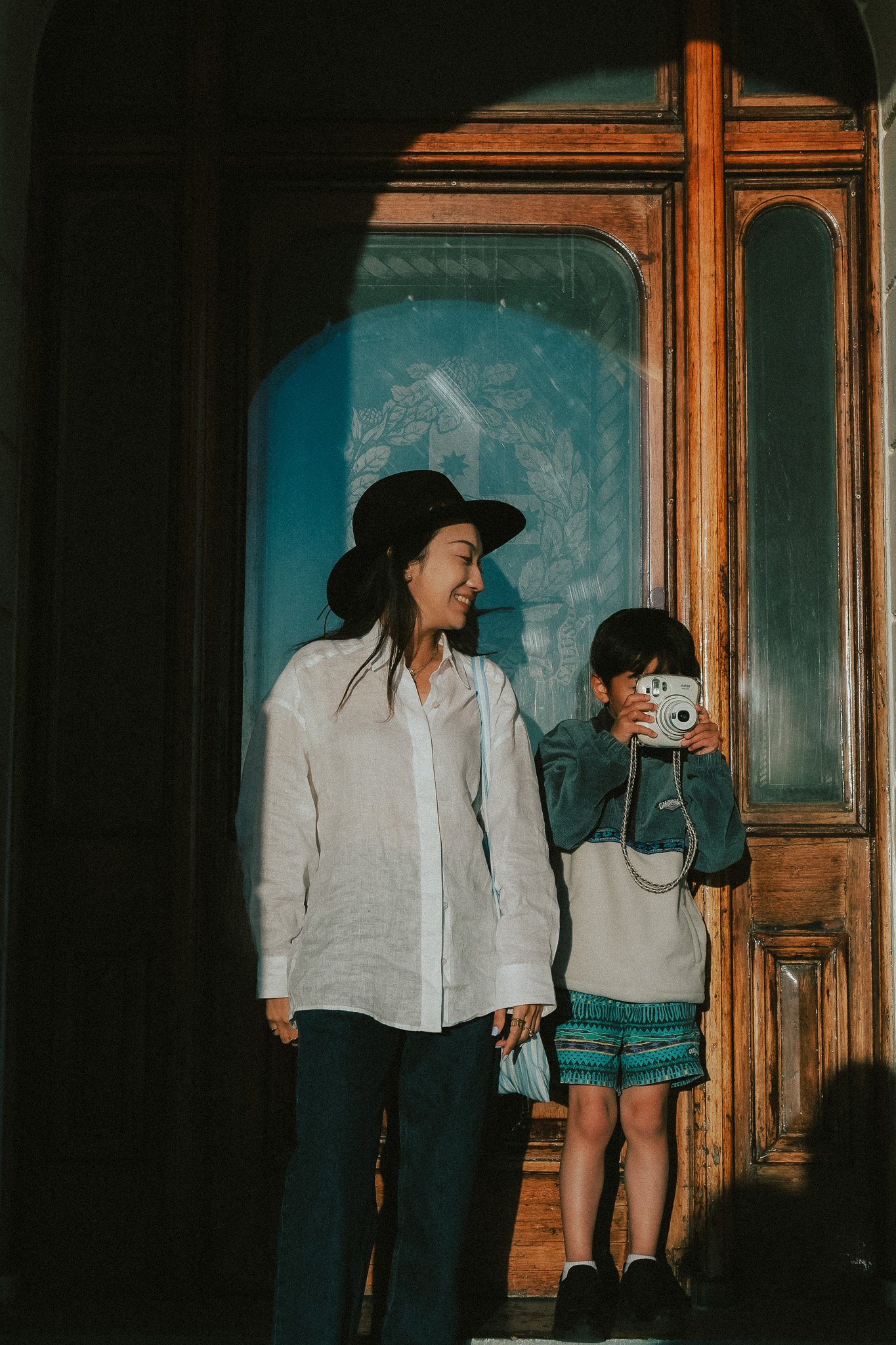 Mum looking down at son beside her as he takes a photo on his Fujifilm Instax, bright afternoon sunlight