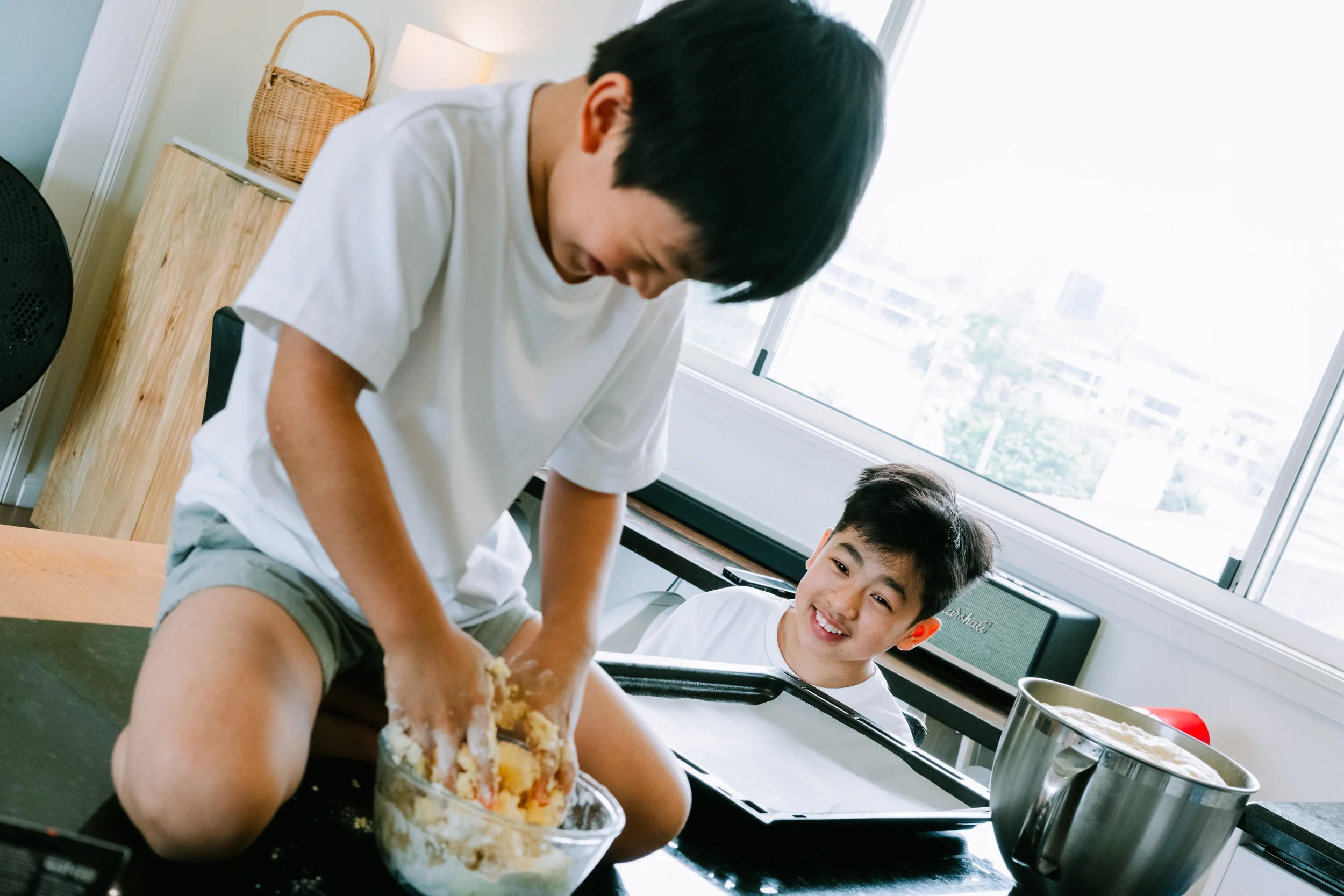 Two boys in the kitchen, one kneading dough on the counter while the other one is watching