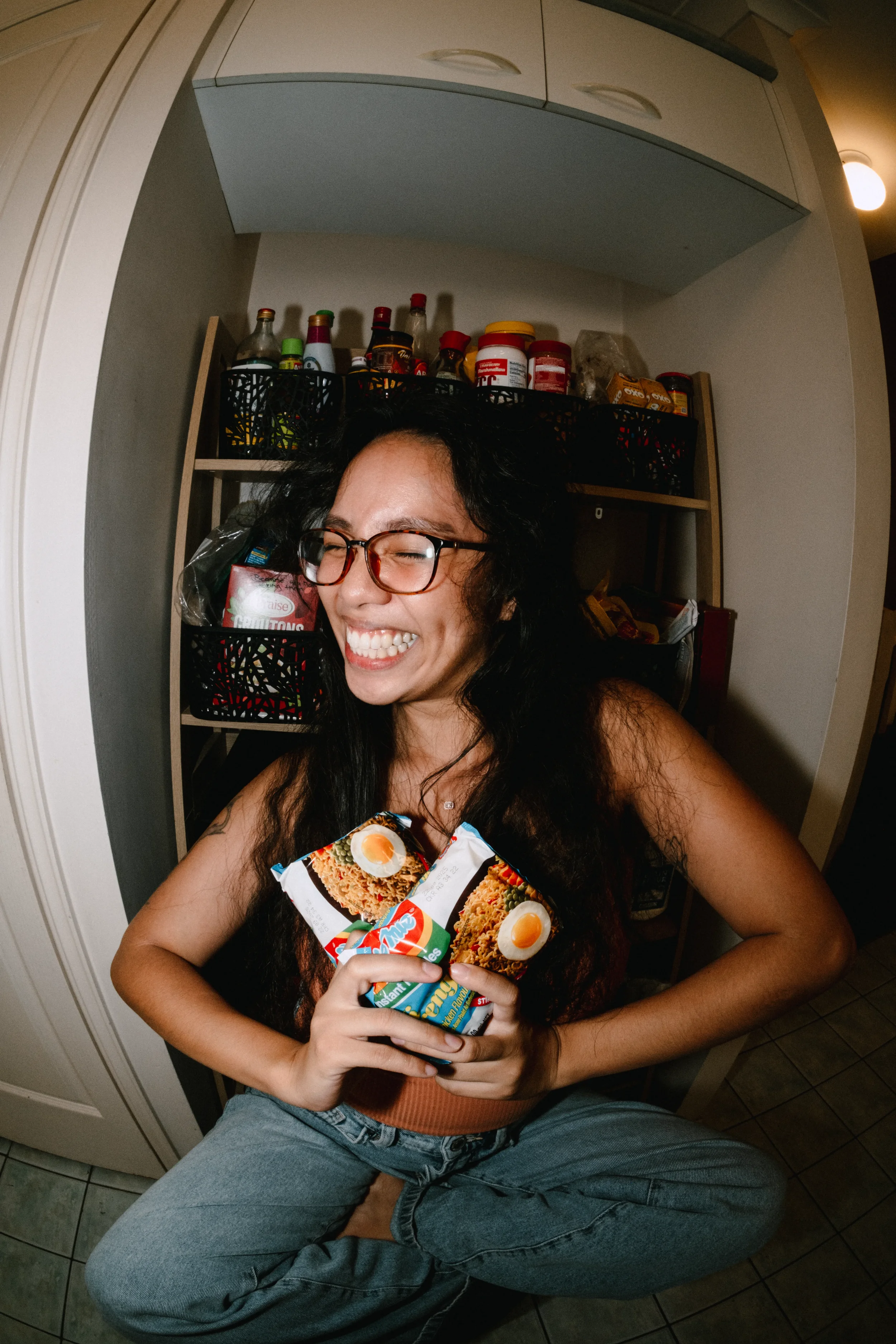 CK, Brand Designer, sitting cross-legged in front of her kitchen pantry, holding packets of Indomie Mee Goreng noodles to her chest