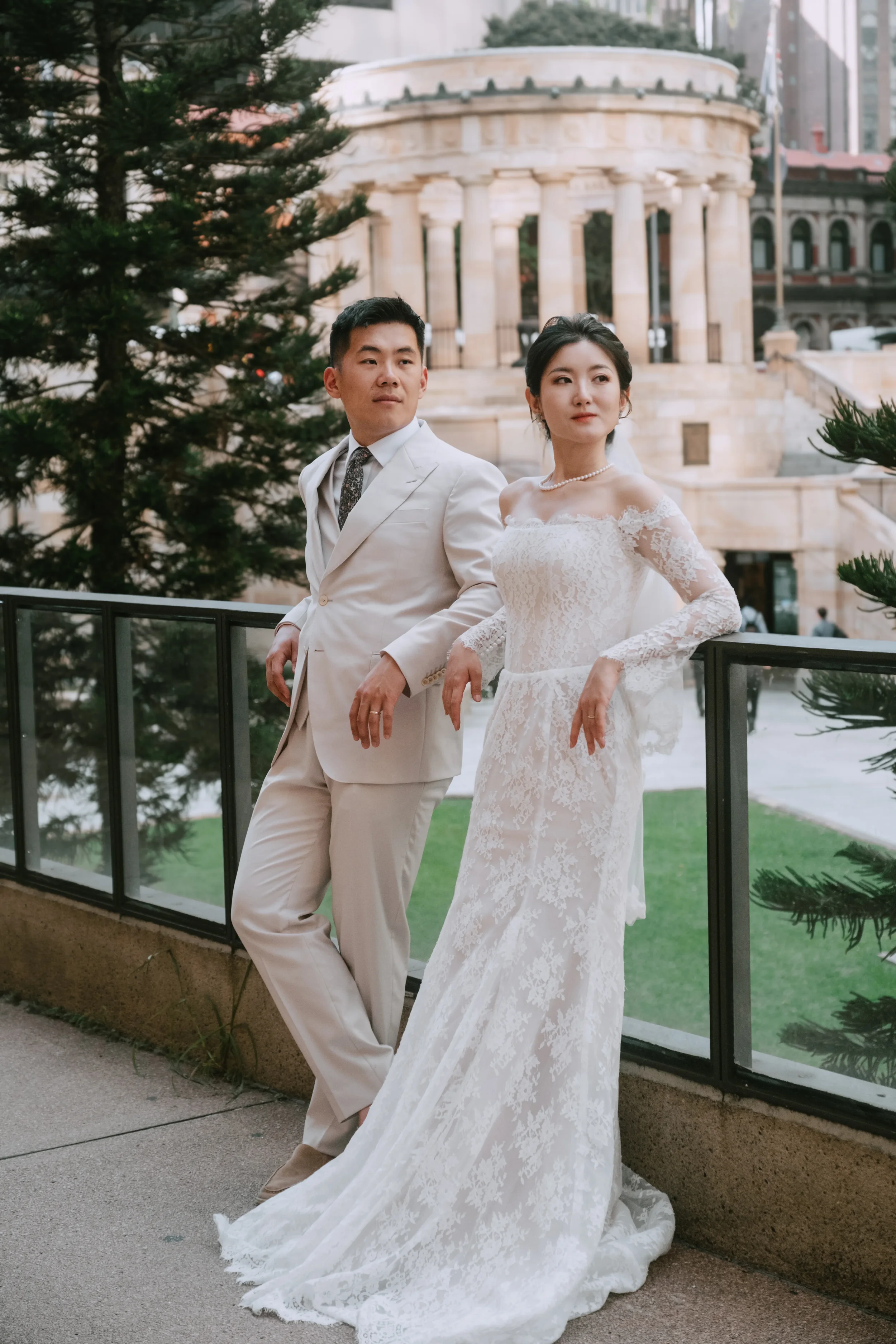 Couple standing side by side, leaning back against glass railings, Anzac Square Memorial Galleries visible behind them