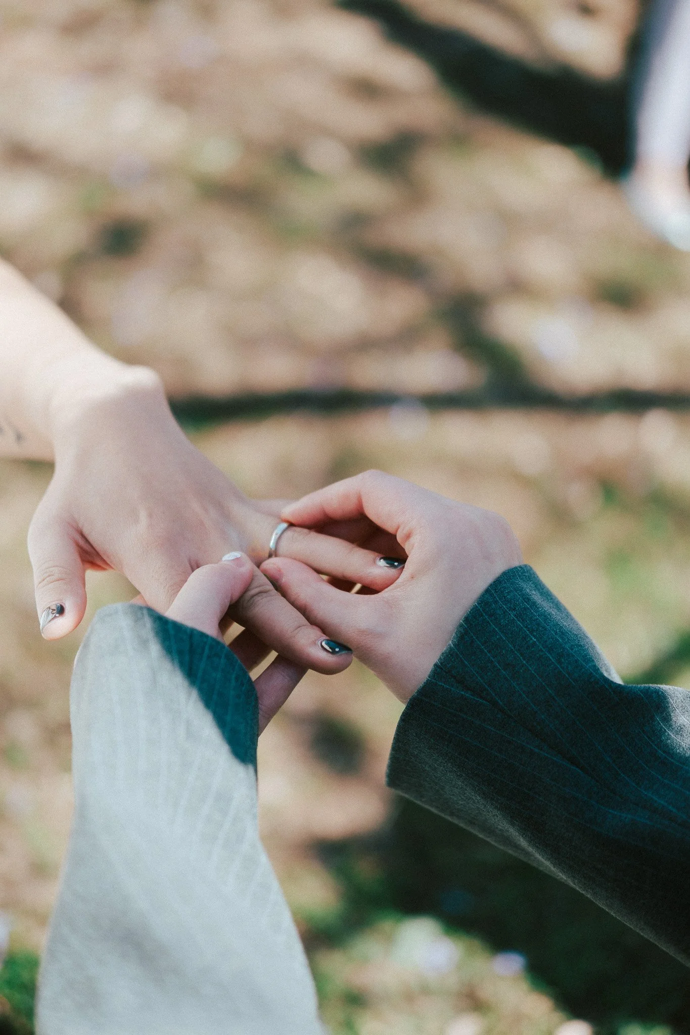Close detail of one partner sliding a wedding band onto the other's ring finger, soft grass visible below