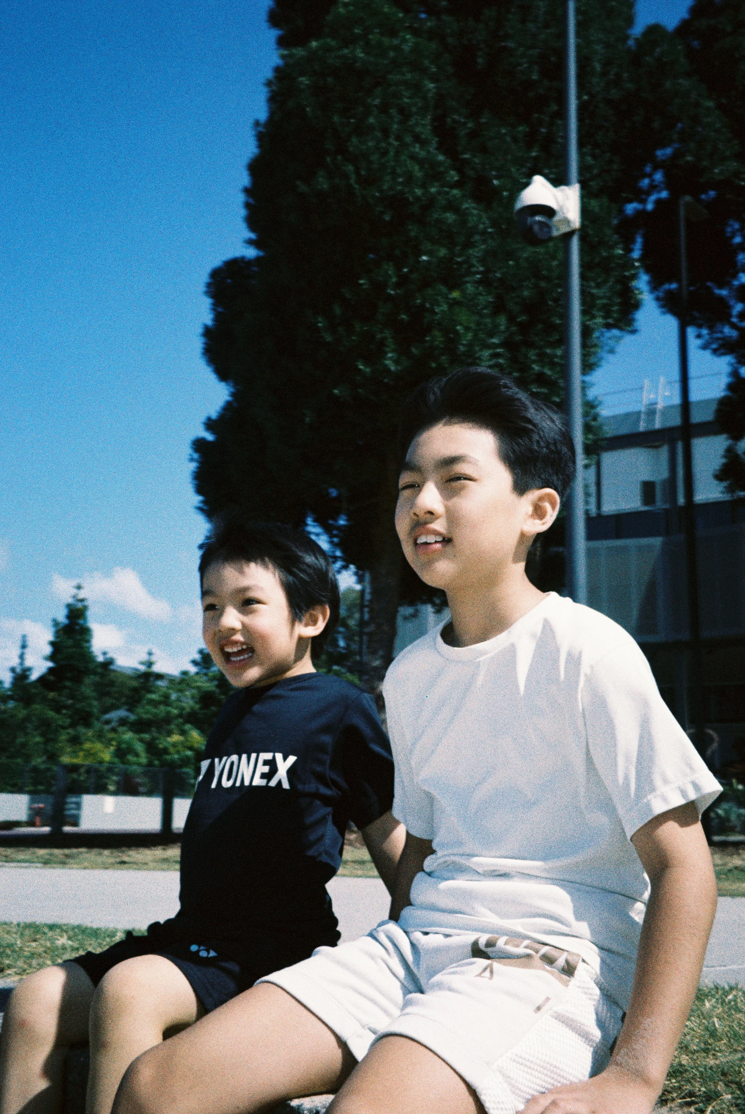 The two boys sitting together on the grass beneath a tree, bright morning light, open sky behind them