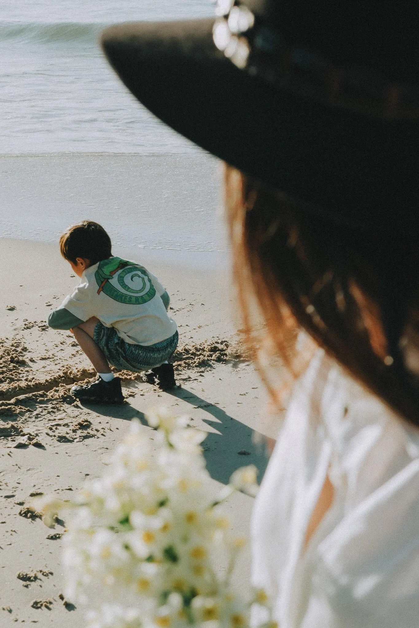 Over Mum's shoulder, her hat visible in the soft foreground, son is playing in the sand