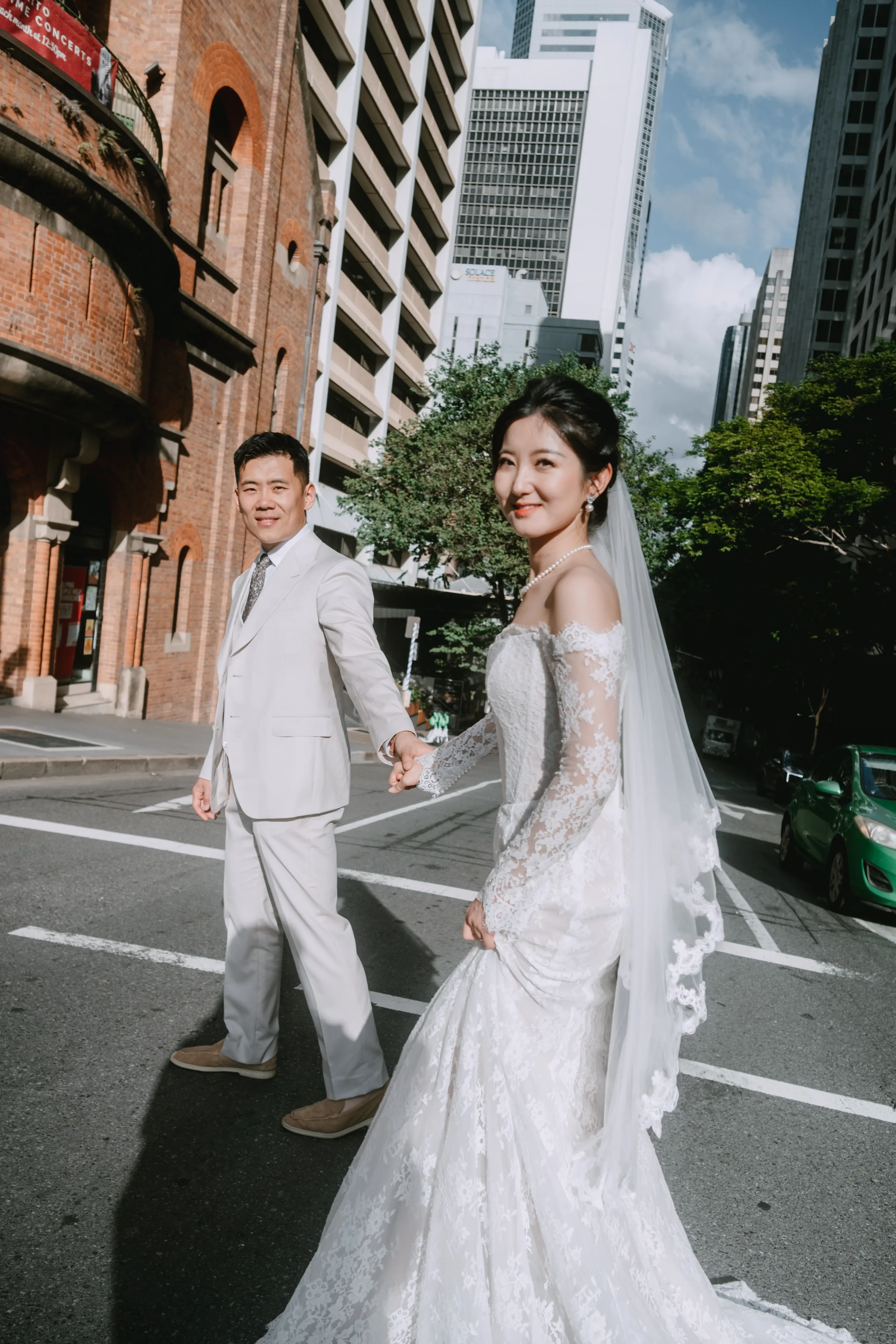 Bridal couple walking hand in hand across a city street, both smiling toward the camera, Brisbane City architecture behind them