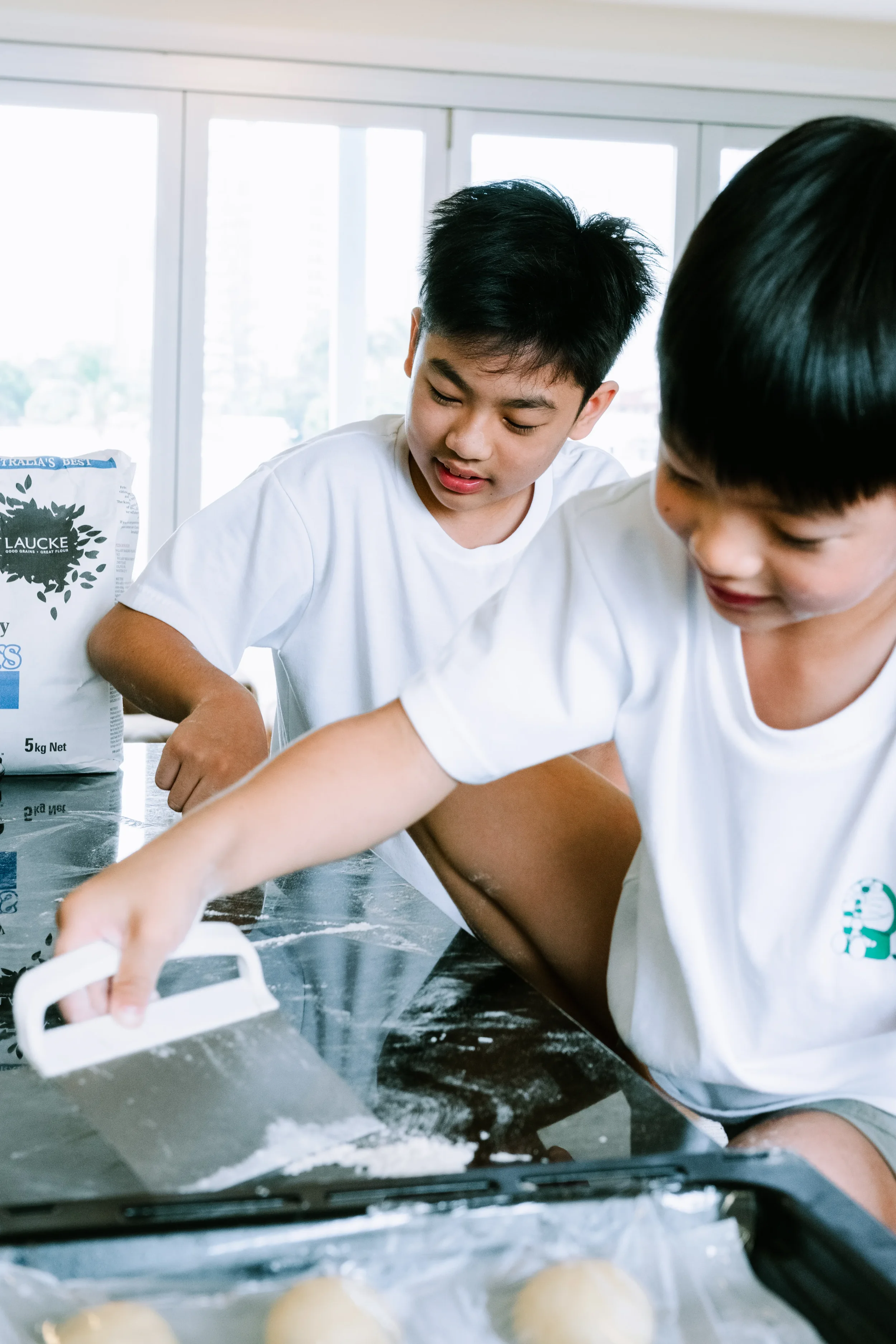 Two boys scraping flour off a dark kitchen counter, bright morning light streaming through the windows behind them