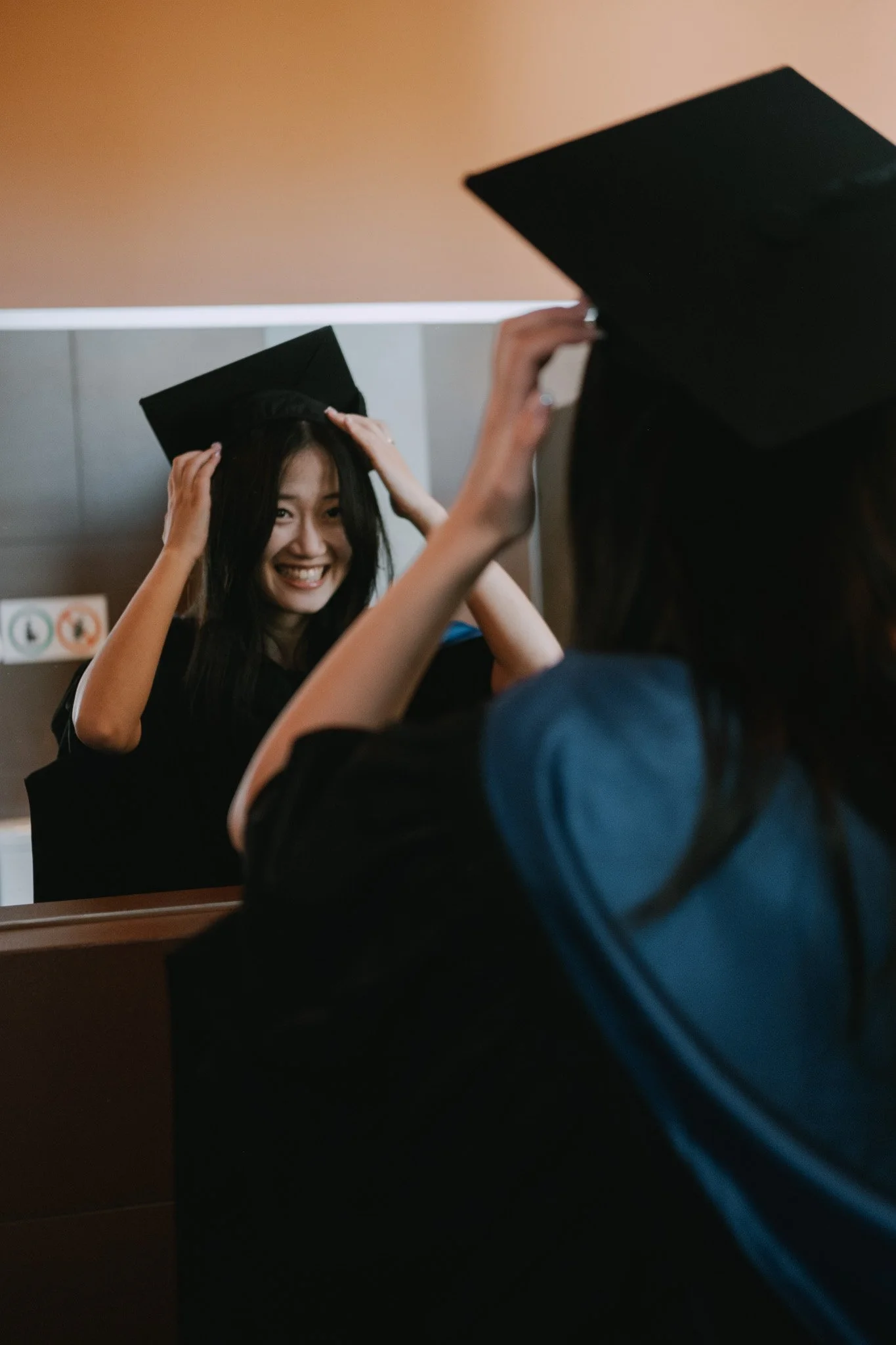 Graduate smiling at her reflection in a mirror as she places her mortarboard on her head