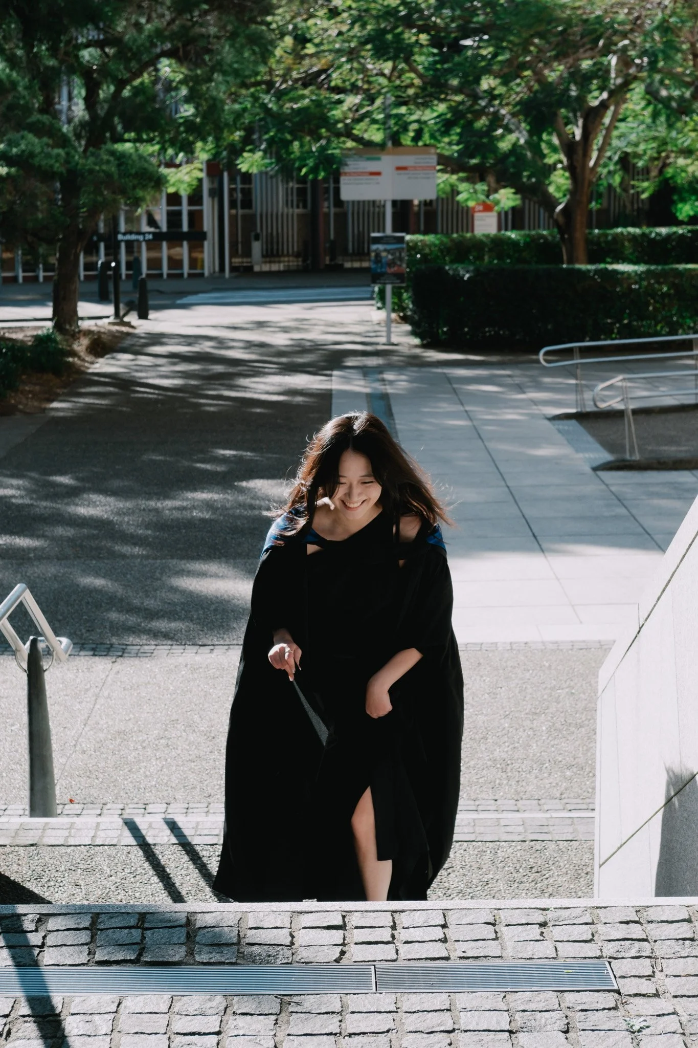Graduate walking up concrete stairs at the University of Queensland, diploma in hand, smiling in bright afternoon light