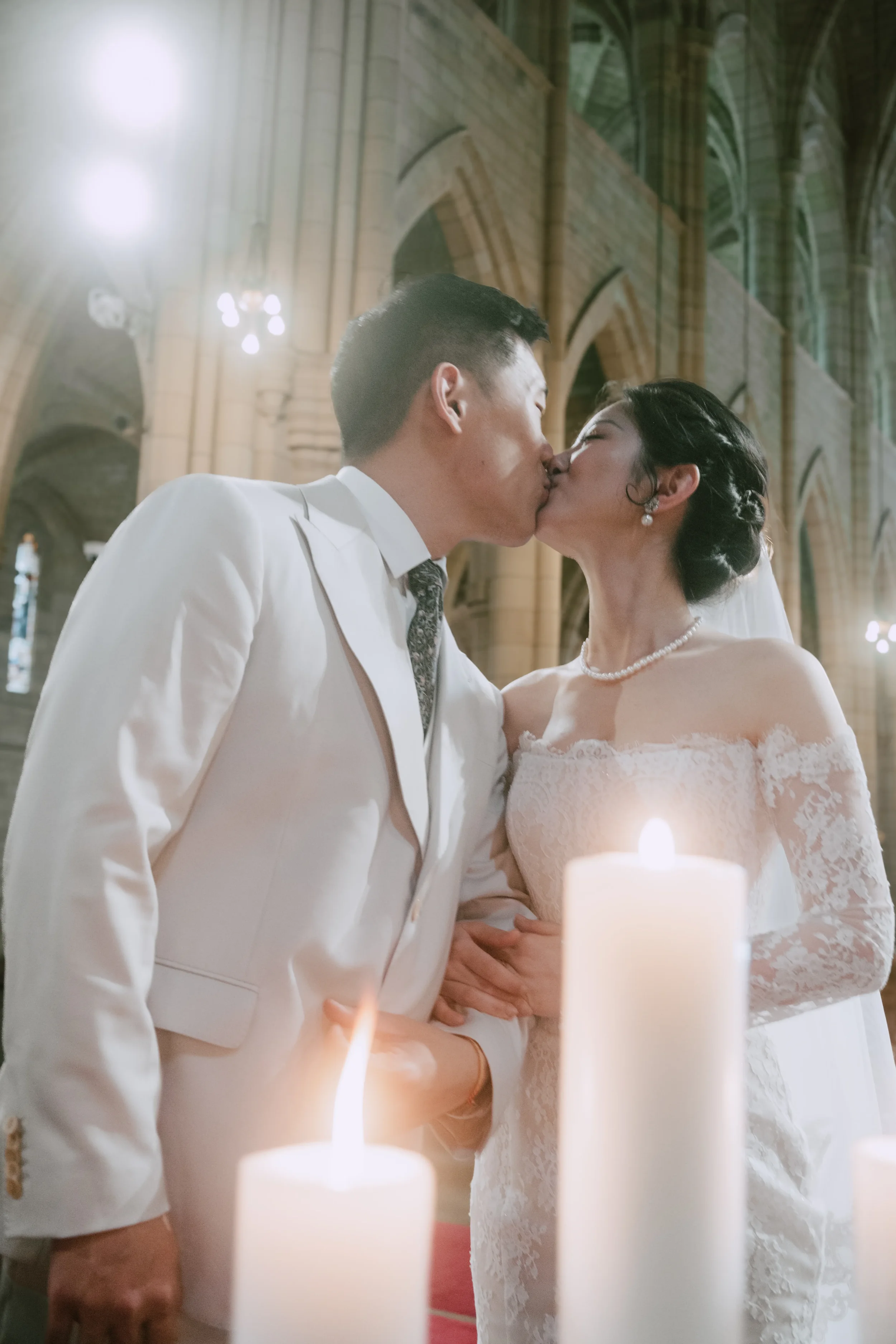 Couple kissing with arms linked, soft candlelight blurred in the foreground, bright chandelier lights behind them