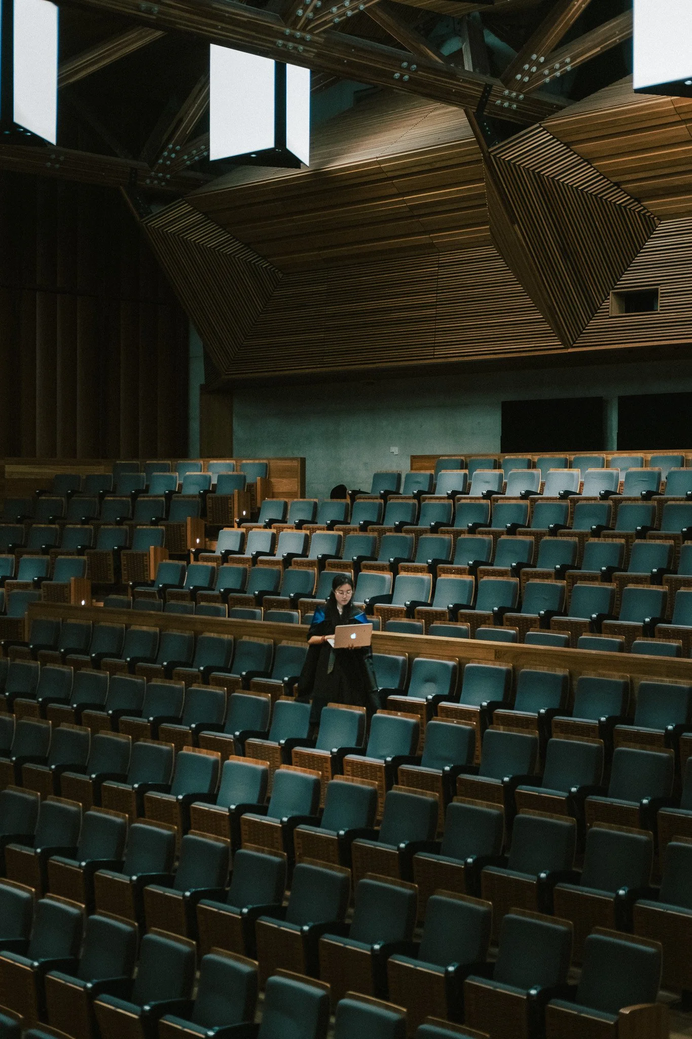 Woman in graduation robes standing alone in a lecture room at the University of Queensland, soft ceiling light above her