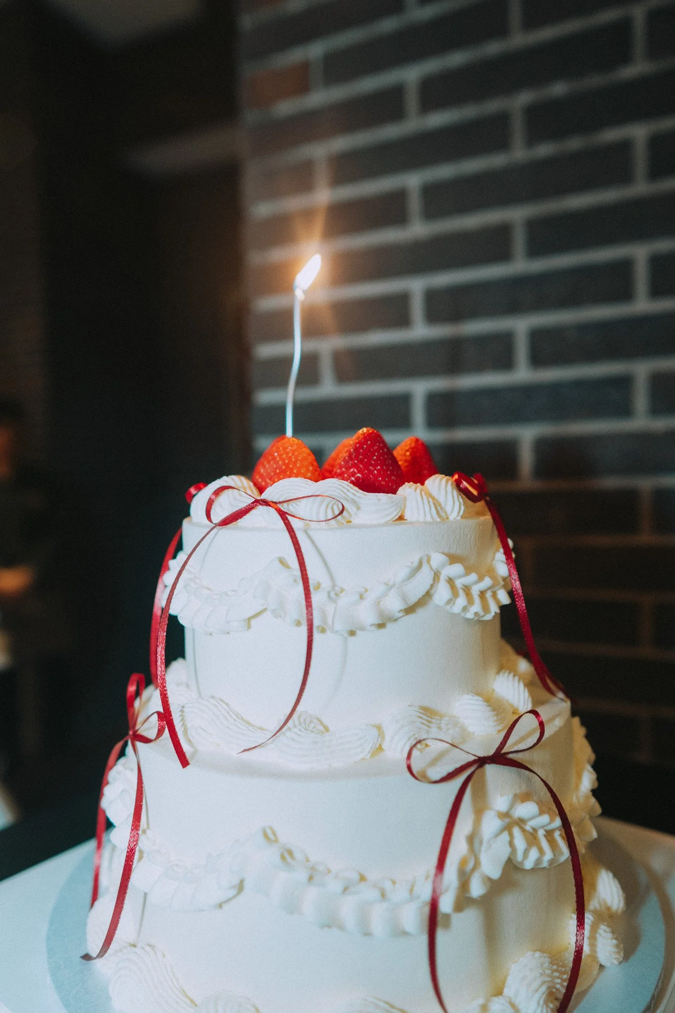 Two-tiered cake in white icing, decorated with thin red ribbons, topped with red strawberries and a candle