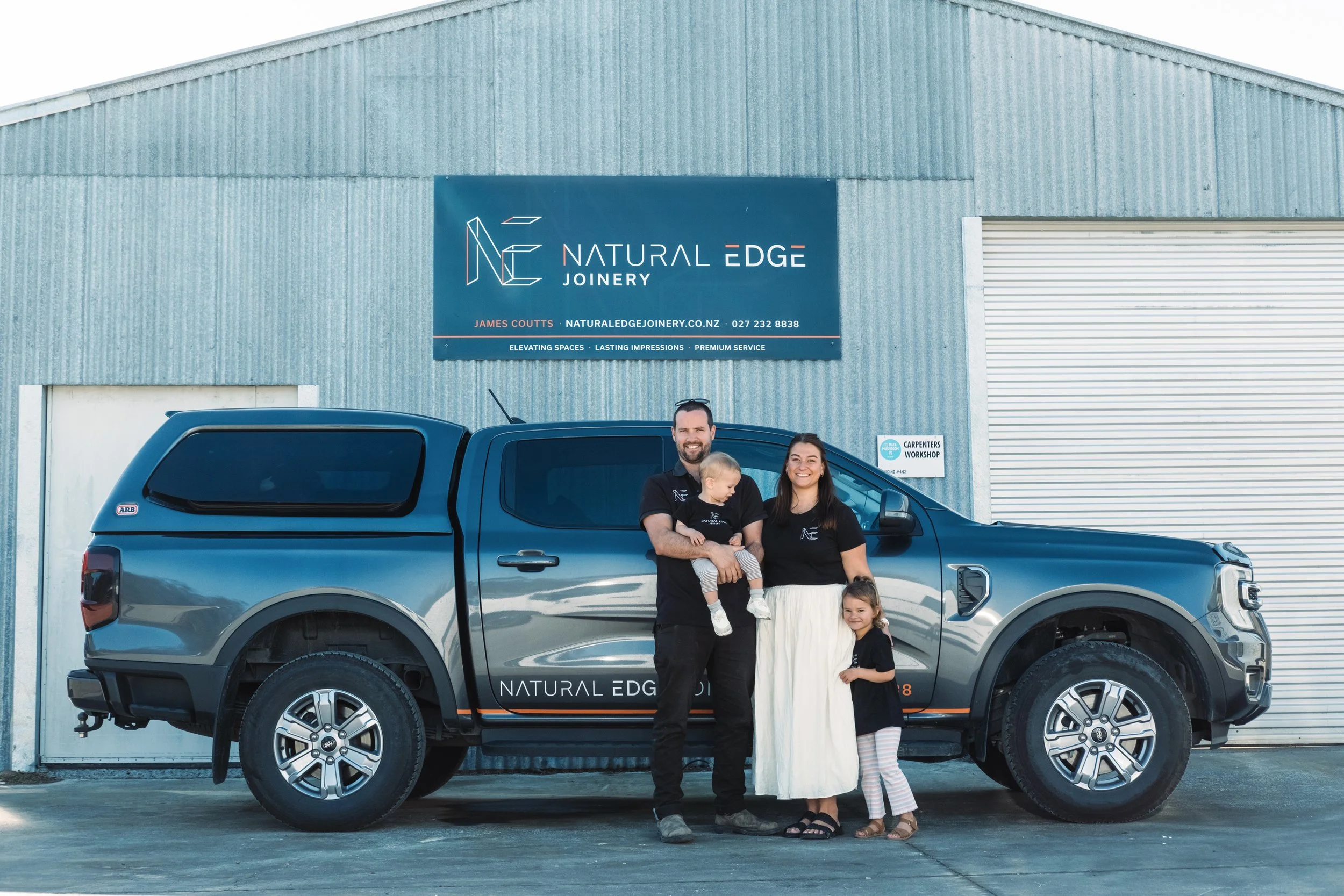 A family of four standing in front of a silver pickup truck parked in front of a blue industrial building with a sign that reads 'Natural Edge Joinery.'