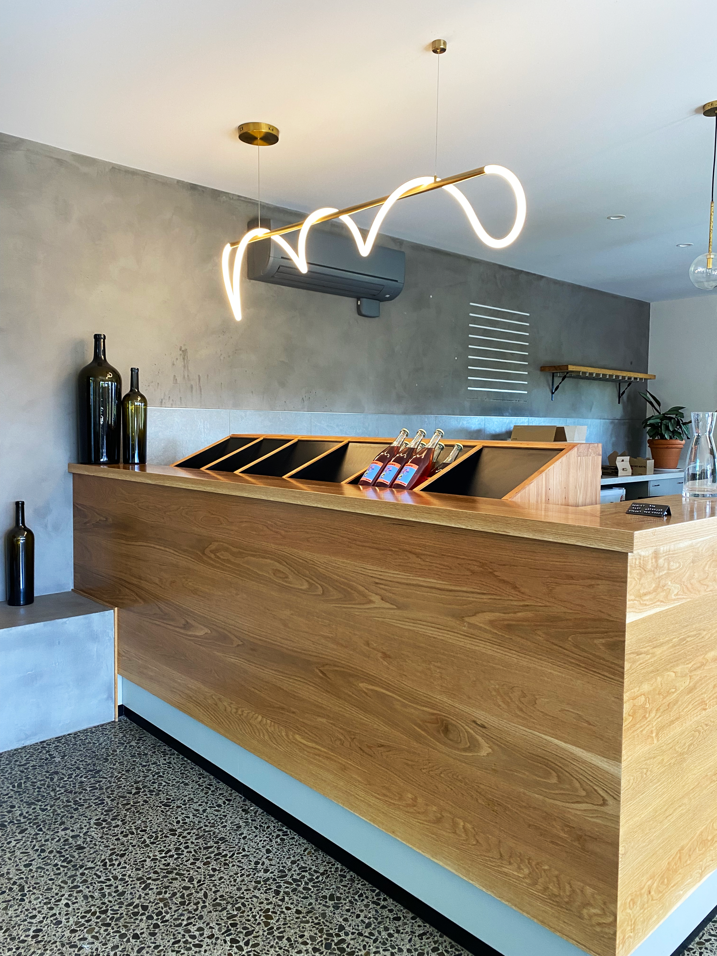 Modern wooden reception counter with three bottles, a stack of check-in papers, a glass water pitcher, and a small plant on the background shelf; contemporary lighting and decor. Custom wine bottle display unit solutions