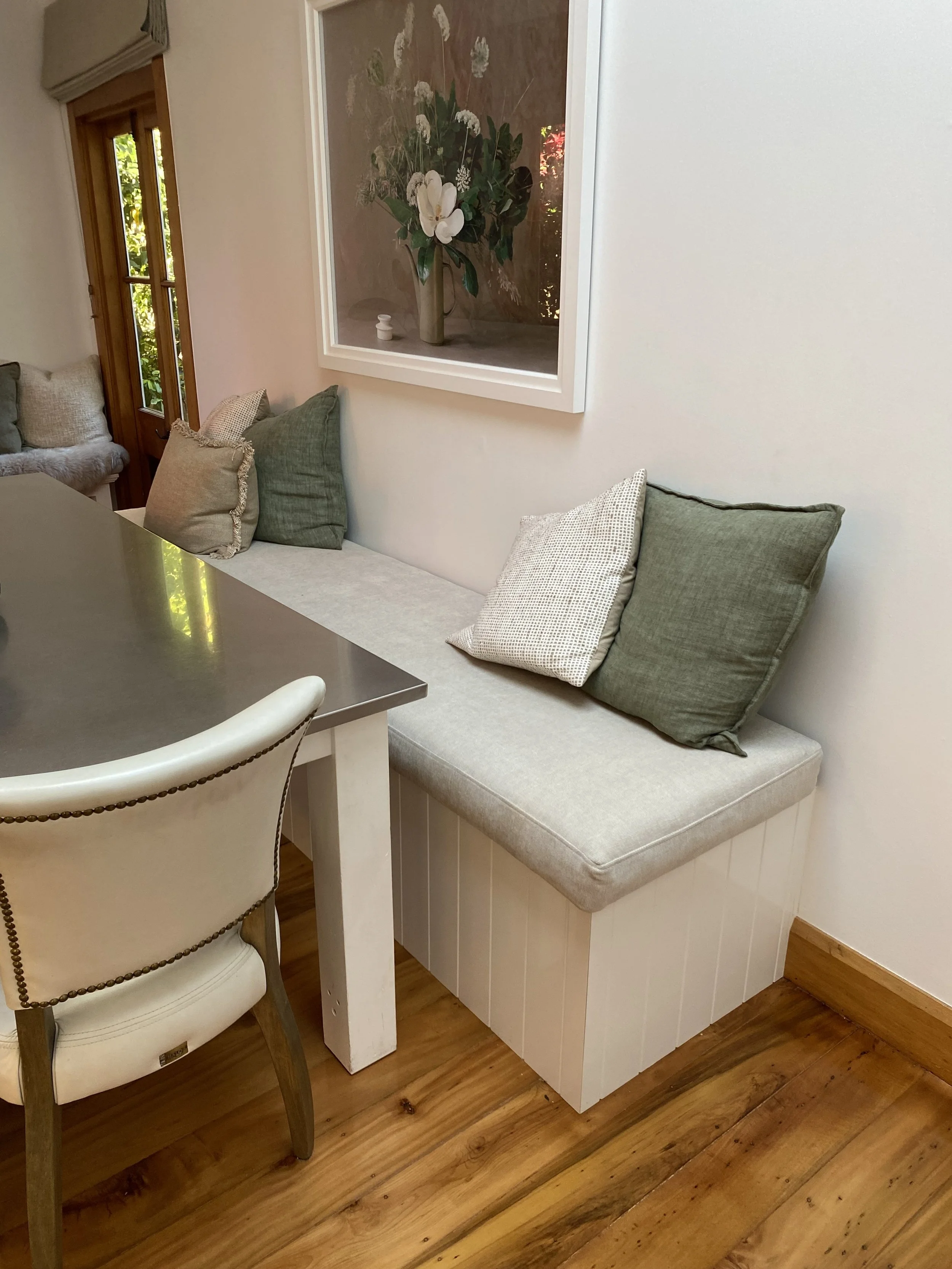 A built-in bench with light gray cushion and four pillows in shades of green, beige, and cream, situated against a white wall in a dining area. Custom comfort bench seat to suit the home and lifestyle