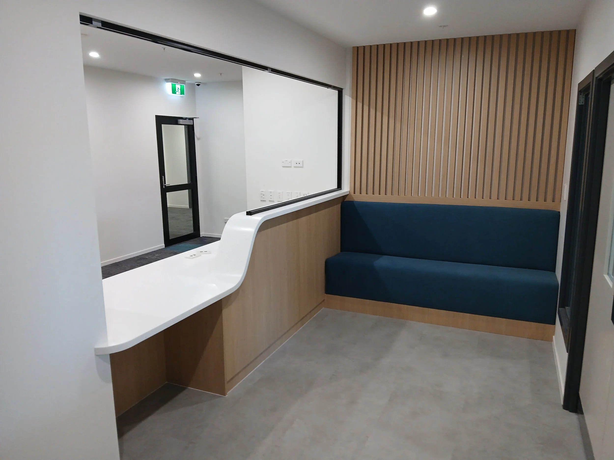 Modern interior reception commercial fit-out, with a white counter with a curve in it, a custom navy blue couch against a wooden slat wall. Cosy and comfort in the waiting room in this reception area